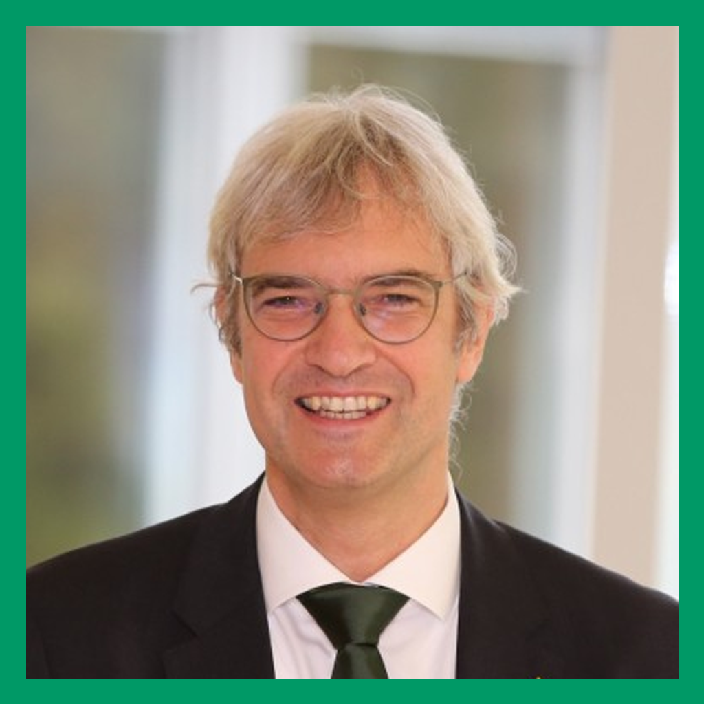 Portrait of Frank Heinze. A grey-haired man in rounded glasses smiles at the camera. He is wearing a black suit jacket over a white shirt, with a dark green necktie.