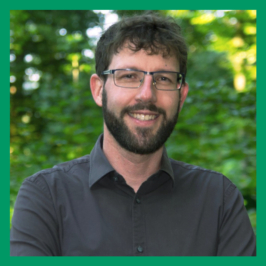 Portrait of Thorsten Reitz. A white man with a neatly trimmed beard and brown hair smiles at the camera. He is wearing square metal glasses and a charcoal button down shirt.