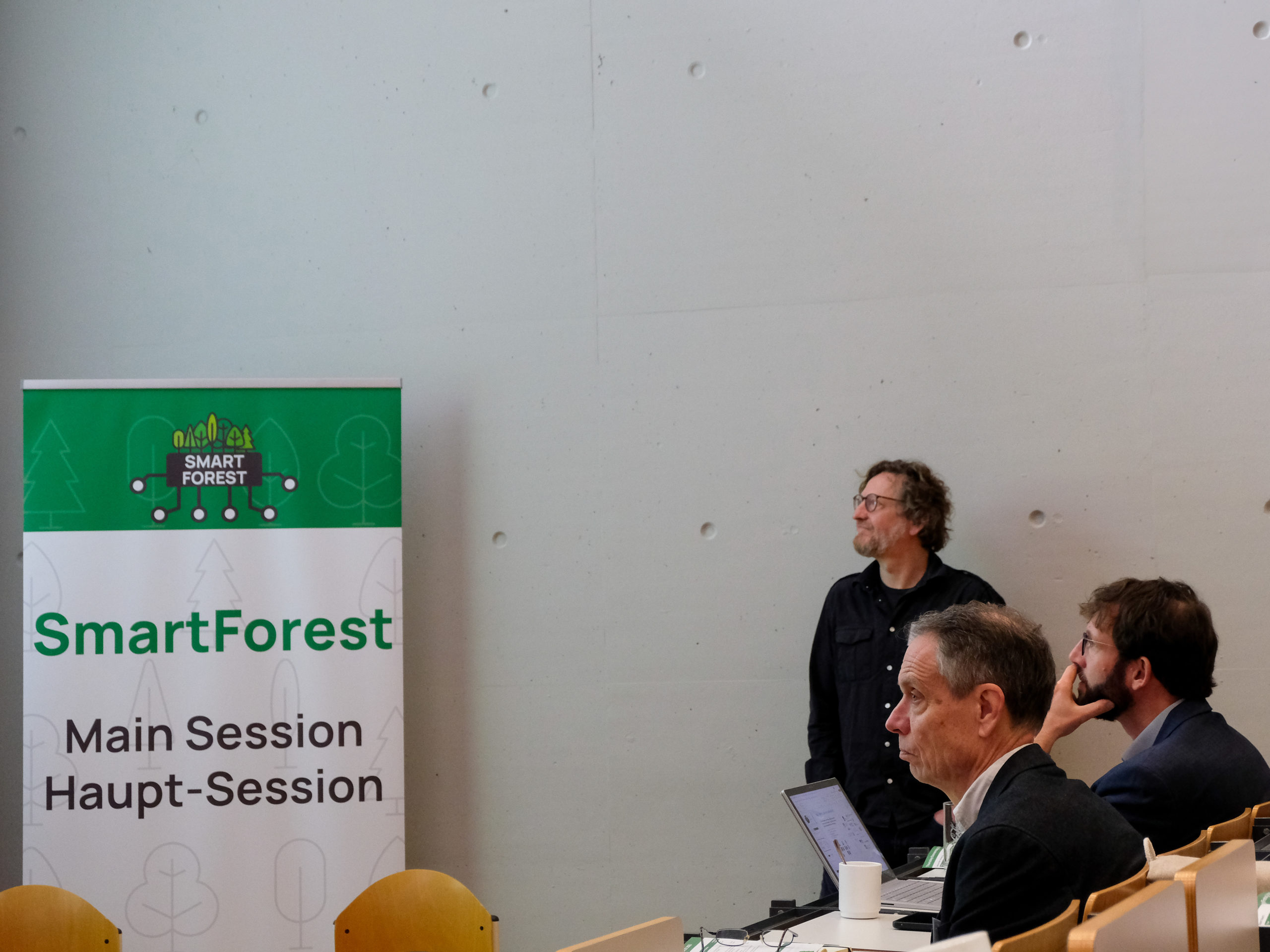 Three men watch the front of the lecture hall with interest. Next to them is a SmartForest-branded sign with the text "Main Session Hauptsession".