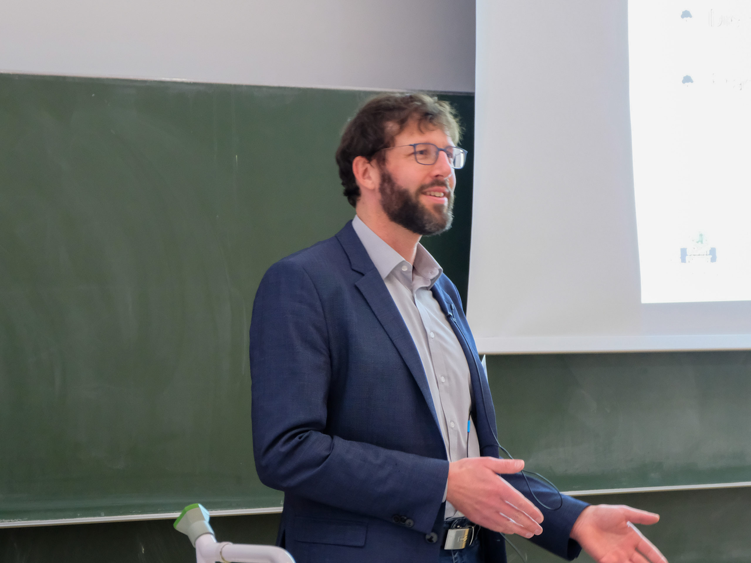Thorsten Reitz, a bespectacled man with brown curly hair, a short beard, a grey shirt, and blue jacket smiles at the crowd as he presents at SmartForest 2025.