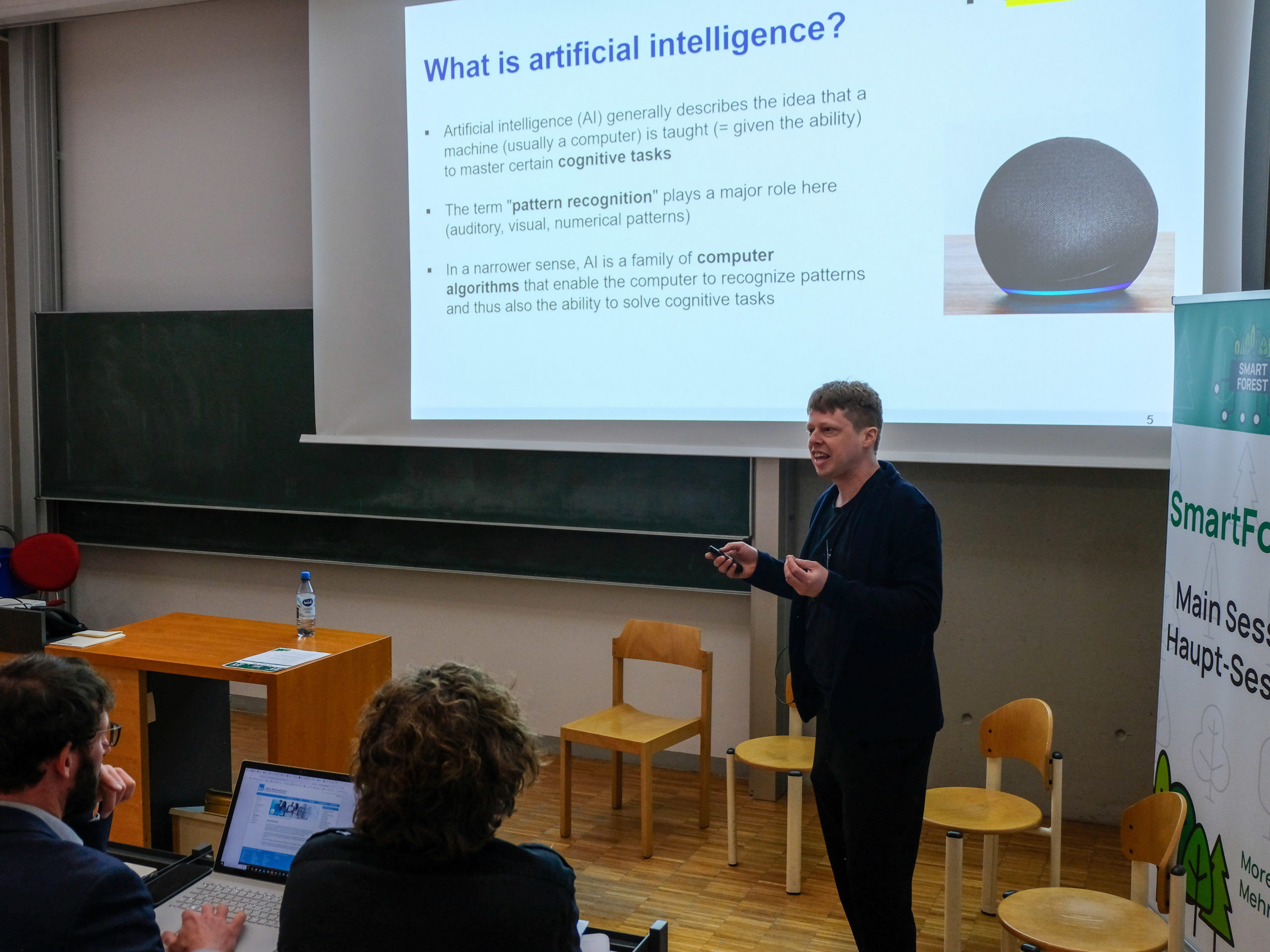 Prof. Dr. Fabian Faßnacht speaks to the crowd in the main lecture hall. Behind him, a slide shows a picture of a digital assistant with the text "What is artificial intelligence?" write large above it. On the slide are three points: "- Artificial Intelligence (AI) generally describes the idea that a machine (usually a computer) is taught (=given the ability) to master cognitive tasks. - The term "pattern recognition" plays a major role here (auditory, visual, numerical patterns). - In a narrower sense, AI is a family of computer algorithms that enable the computer to recognize patterns and thus also the ability to solve cognitive tasks."