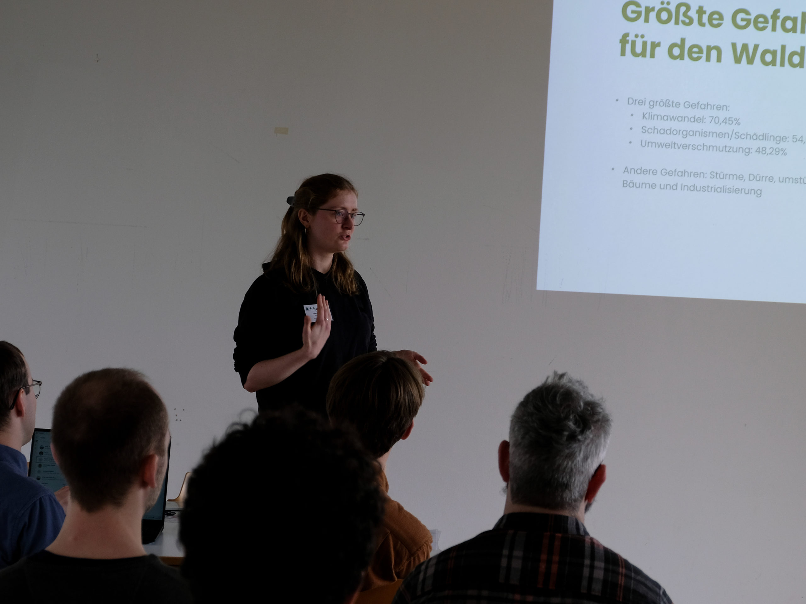 A blonde woman in glasses stands in front of a classroom, gesturing as she addresses the crowd. To the right, a projected slide titled "Größte Gefahren für den Wald" is partially visible, listing the three main dangers to the forest as "Klimawandel: 70.45%, Schadenorganismen/Schädlinge: 54.54%, Umweltverschmutzung: 48.29%". Another point collects other dangers: "Stürme, Dürre, umstürzende Bäume und Industrialisierung".