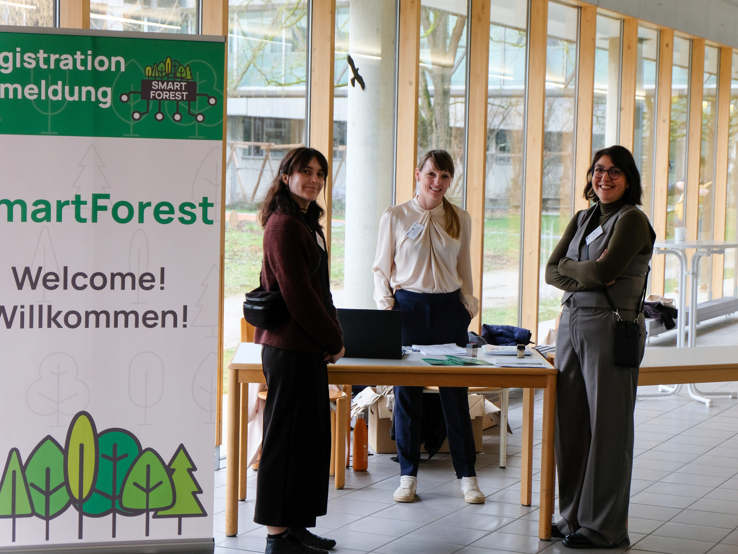 Three women, Ramona Resch, Franziska Hochenegger, and Dr. Nina Krüger, smile at the camera from the SmartForest reception desk.