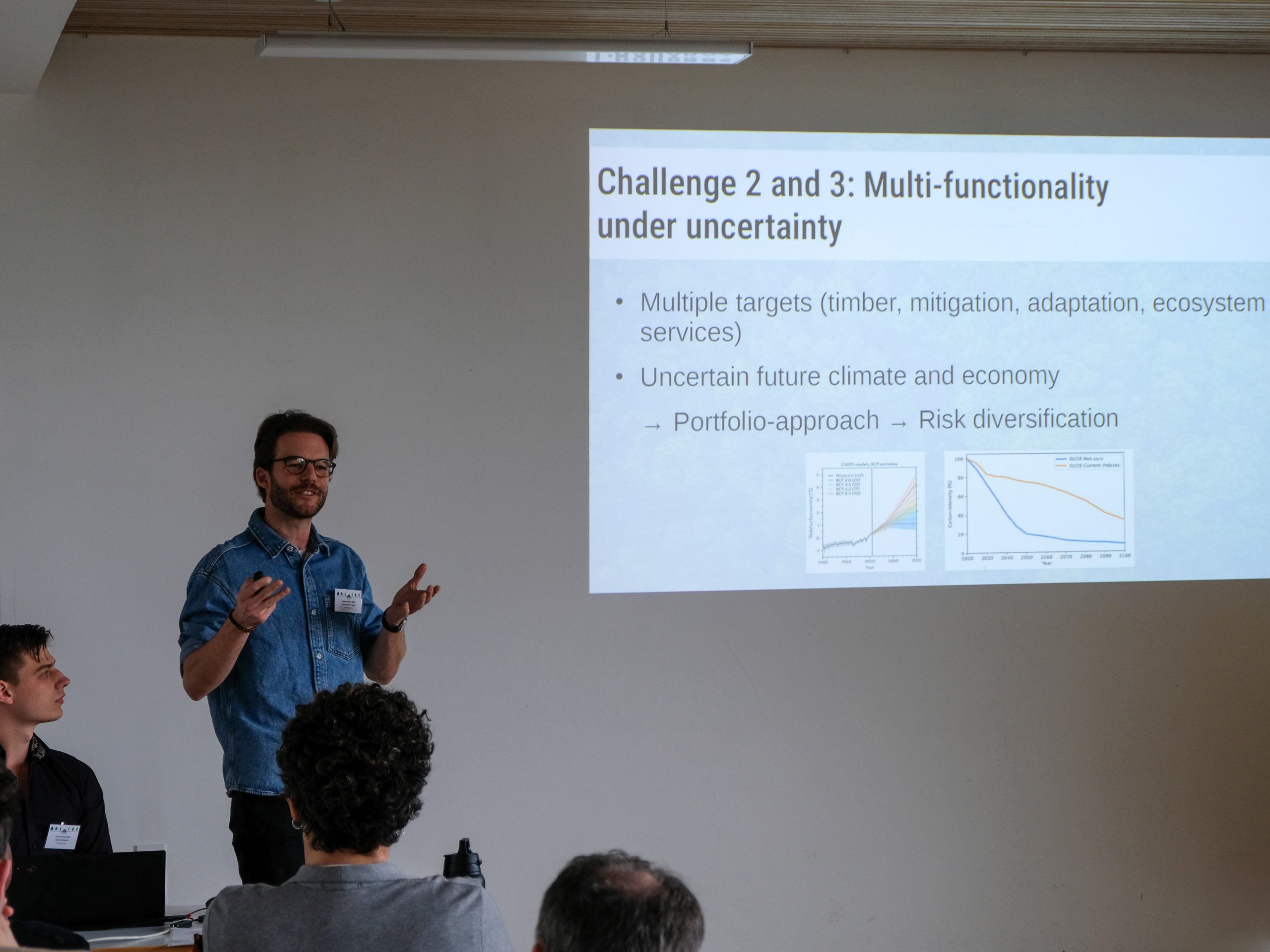 A dark-haired, bespectacled man in a denim shirt stands in front of a classroom, explaining something. Next to him is a projected slide, titled "Challenge 2 and 3: Multi-functionality under uncertainty" It has two points: "Multiple targets (timber, mitigation, adaptation, ecosystem services" and "Uncertain future climate and economy → Portfolio-approach → Risk diversification", with two graphs along the bottom.
