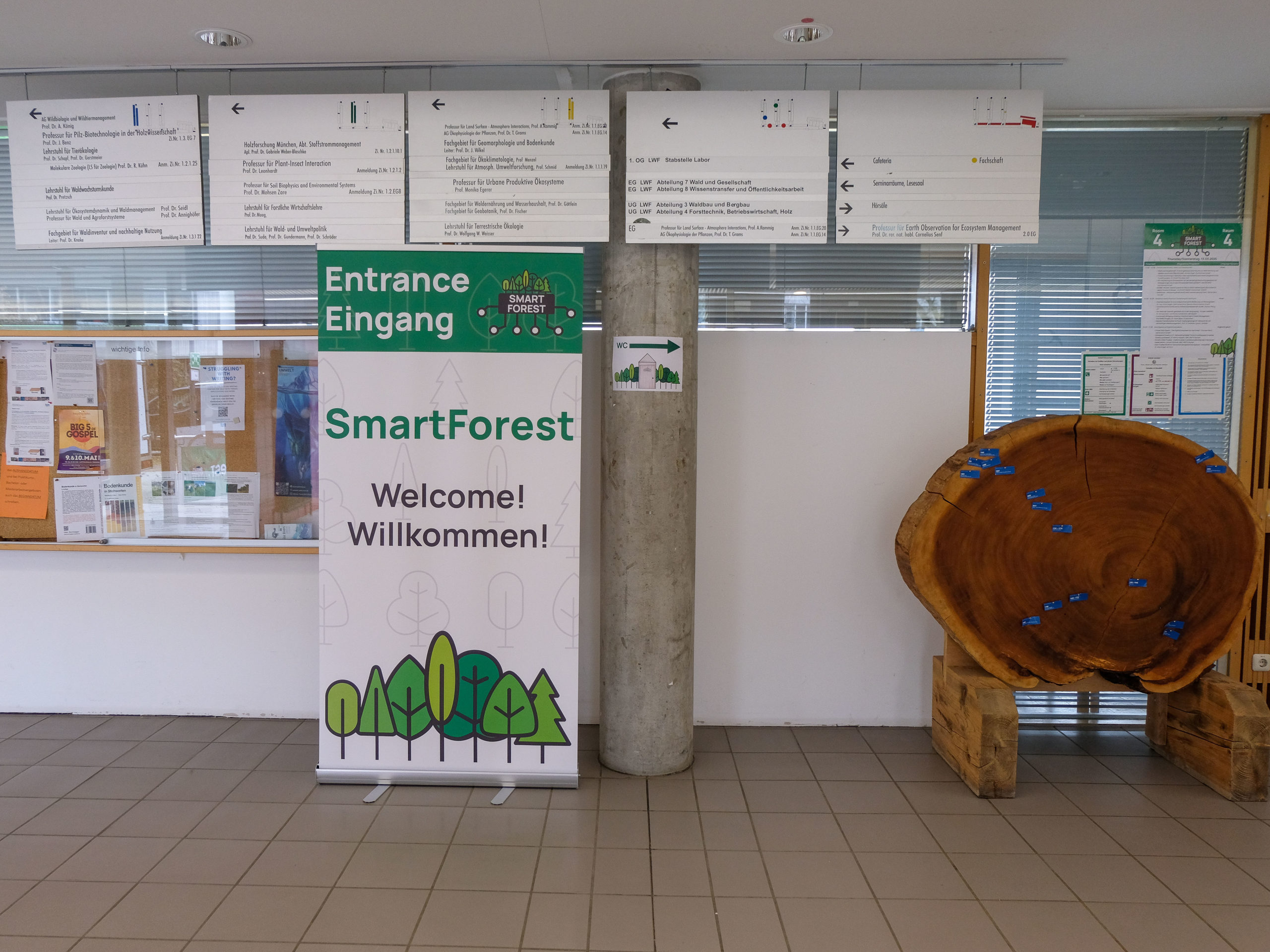 The welcome-sign for SmartForest 2025. Also visible are the building's directional signs overhead, the time schedule for Room 4, a large tree slice used as a pinboard, and a SmartForest sign pointing to the toilets.
