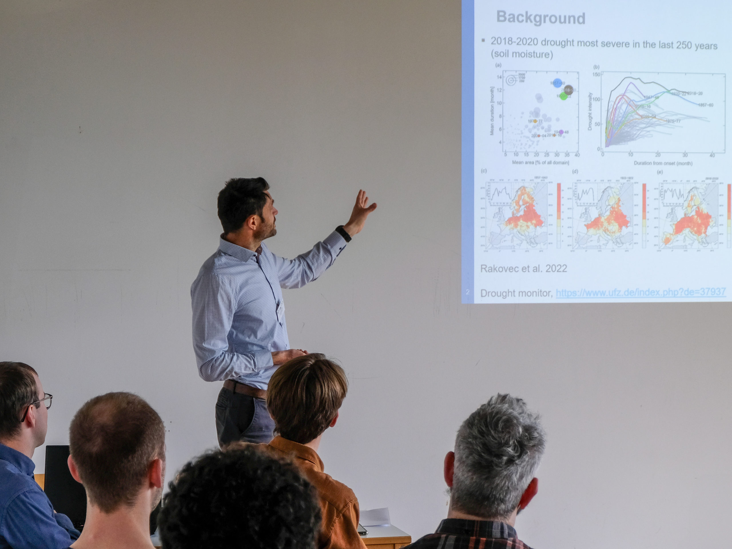 Dr. Frank Thonfeld, a dark-haired man with a stubbly beard and light blue button-up shirt, gestures at a projected slide titled "Background" as a classroom of interested people looks on. The slide reads "2018-2020 drought most severe in the last 250 years (soil moisture)" and shows several graphs and maps of Europe with location data. The final two lines are "Rakovec et al. 2022" and "Drought monitor", with a link to the UFZ website.