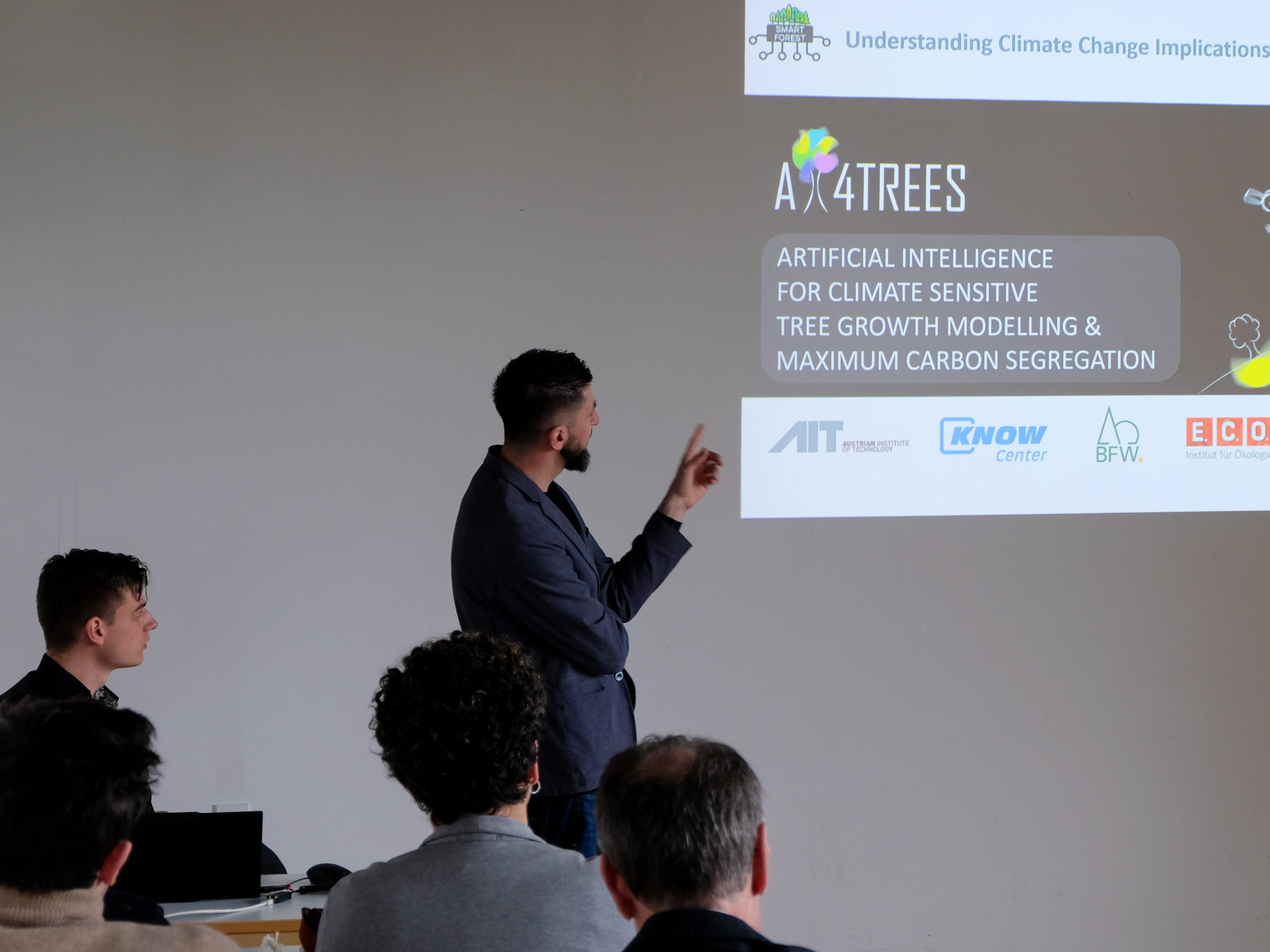 Refiz Duro, a dark-haired man with a beard wearing a blue blazer, points at a slide in front of a classroom. The top of the slide sports the SmartForest logo and the text "Understanding Climate Change Implications". Below is the AI4Trees Project's logo, alongside the text "Artificial Intelligence for Climate Sensitive Tree Growth Modelling & Maximum Carbon Segregation". Below are the logos of the Austrian Institute of Technology, Know Center, BFW, and E.C.O. Institut für Ökologie.
