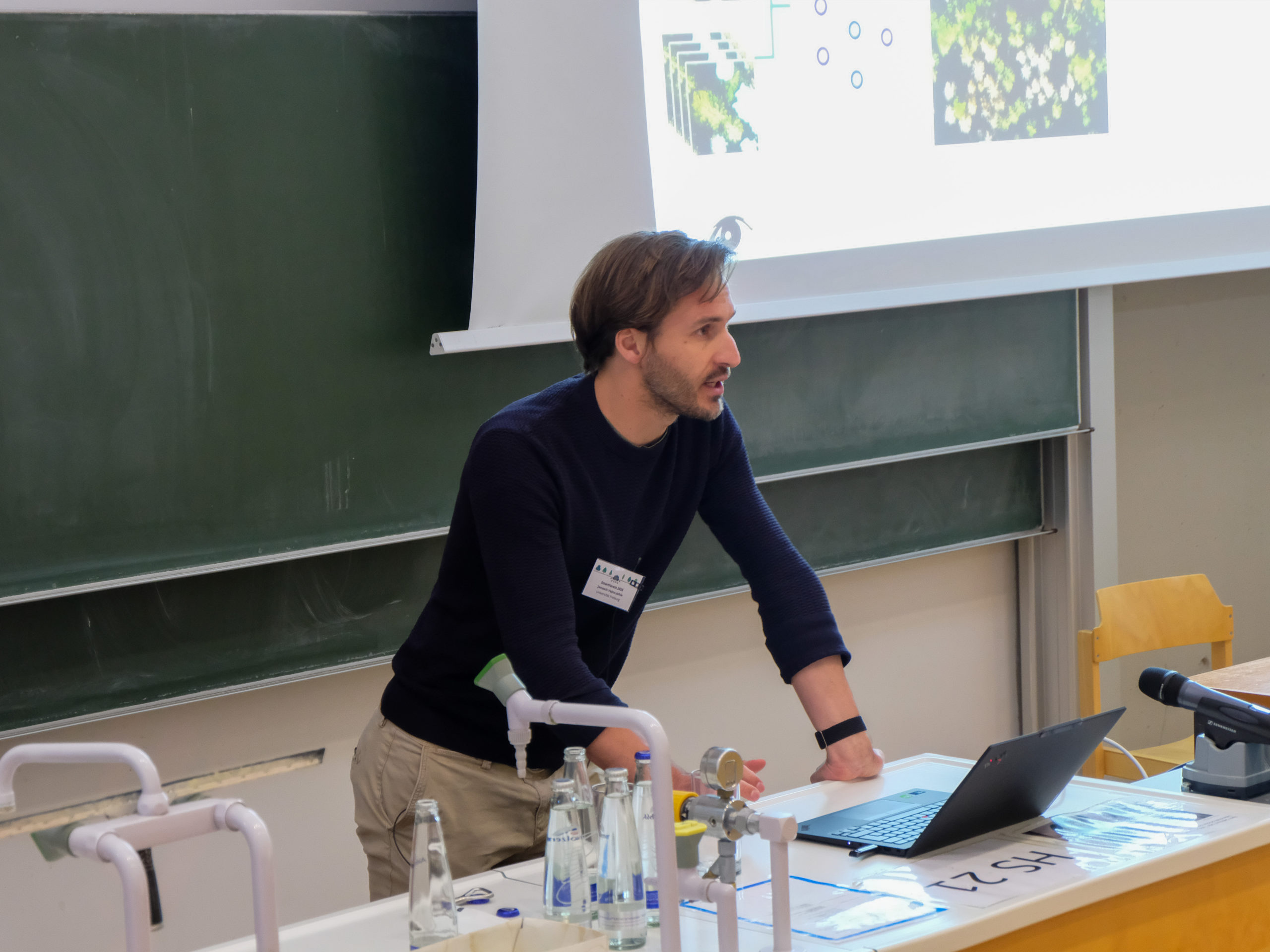 Janusch Vajna-Jehle, a brown-haired man with a short stubbly beard, a dark blue jumper, and tan trousers, leans over a desk as he speaks in the main lecture hall.