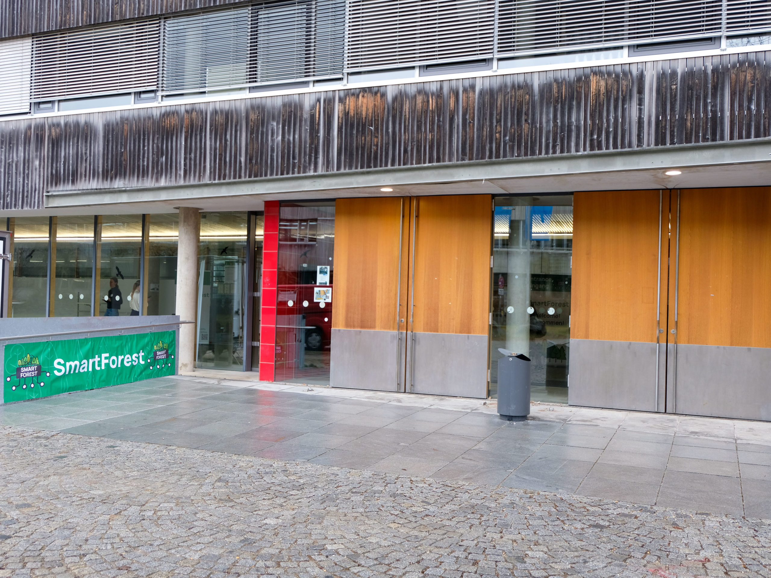 An outdoor shot of the entrance to the 2025 SmartForest conference in Freising, showing two sets of large double doors, as well as an accessibility-friendly automatic sliding door, and a green SmartForest banner.