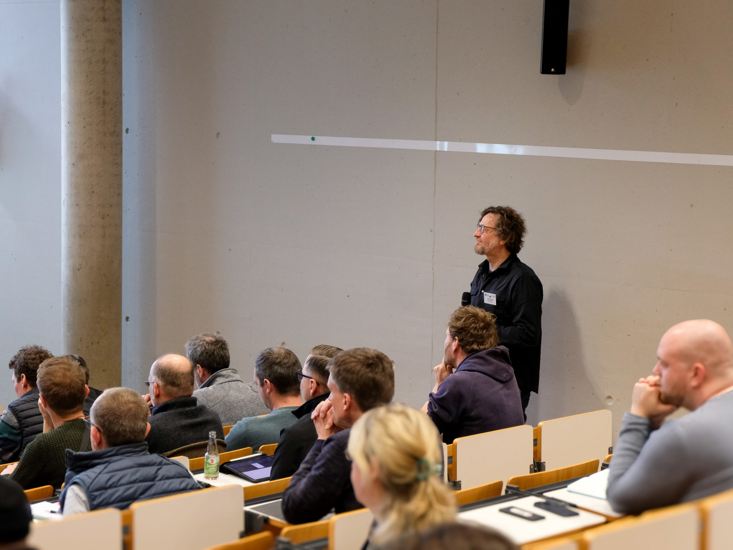Dr. Werner Rammer, a bearded man with glasses and curly brown hair, stands to the side of the well-filled lecture hall during SmartForest, holding a microphone to facilitate crowd questions.
