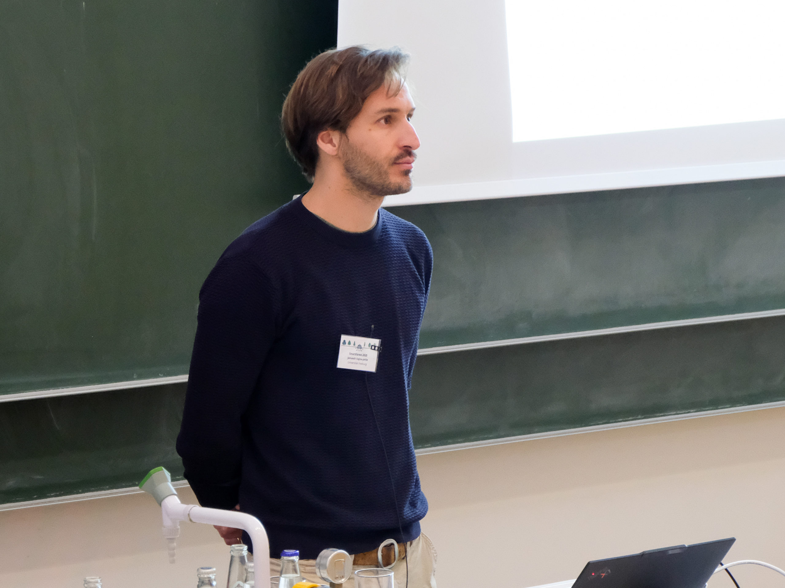 Janusch Vajna-Jehle, a brown-haired man with a stubbly beard and a blue jumper, stands in front of a blackboard with his hands behind his back.