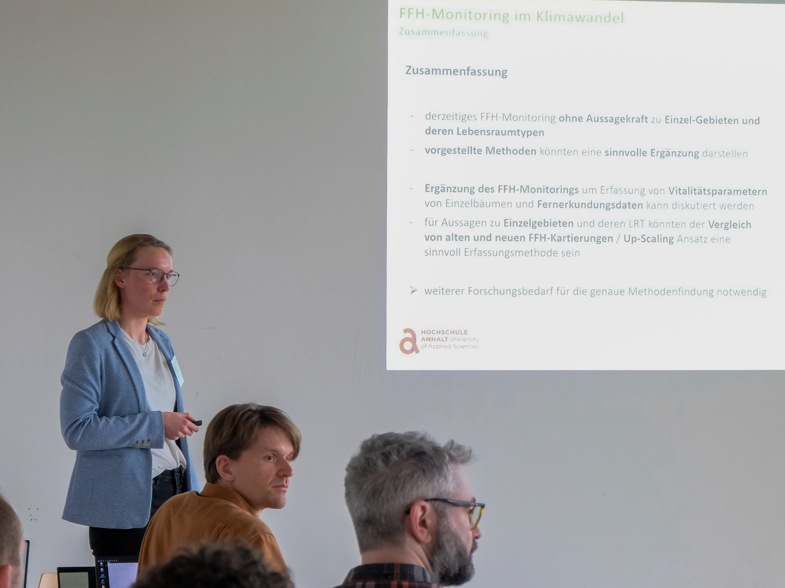 A bespectacled blonde woman in a white shirt and blue blazer is standing in front of a classroom. She and other attendees are attentively looking over at someone out of frame. To the right, a projected slide reads "FFH-Monitoring im Klimawandel. Zusammenfassung.", with the points "-derzeitiges FFH-Monitoring ohne Aussagekraft zu Einzel-Gebieten und deren Lebensraumtypen. - vorgestellte Methoden Könnten eine sinvolle Ergänzung darstellen. - Ergänzung des FFH-Monitorings um Erfassung von Vitalitätsparametern von Einzelbäumen und Fernerkundungsdaten kann diskutiert werden. - für Aussagen zu Einzelgebieten und deren LRT könnten der Vergleich von alten und neuen FFH-Kartierungen / Up-Scaling Ansatz eine sinvoll Erfassungsmethode sein. - weiterer Forschungsbedarf für die genaue Methodenfundung notwendig". At the bottom is the logo of the Hochschule Anhalt University of Applied Sciences.