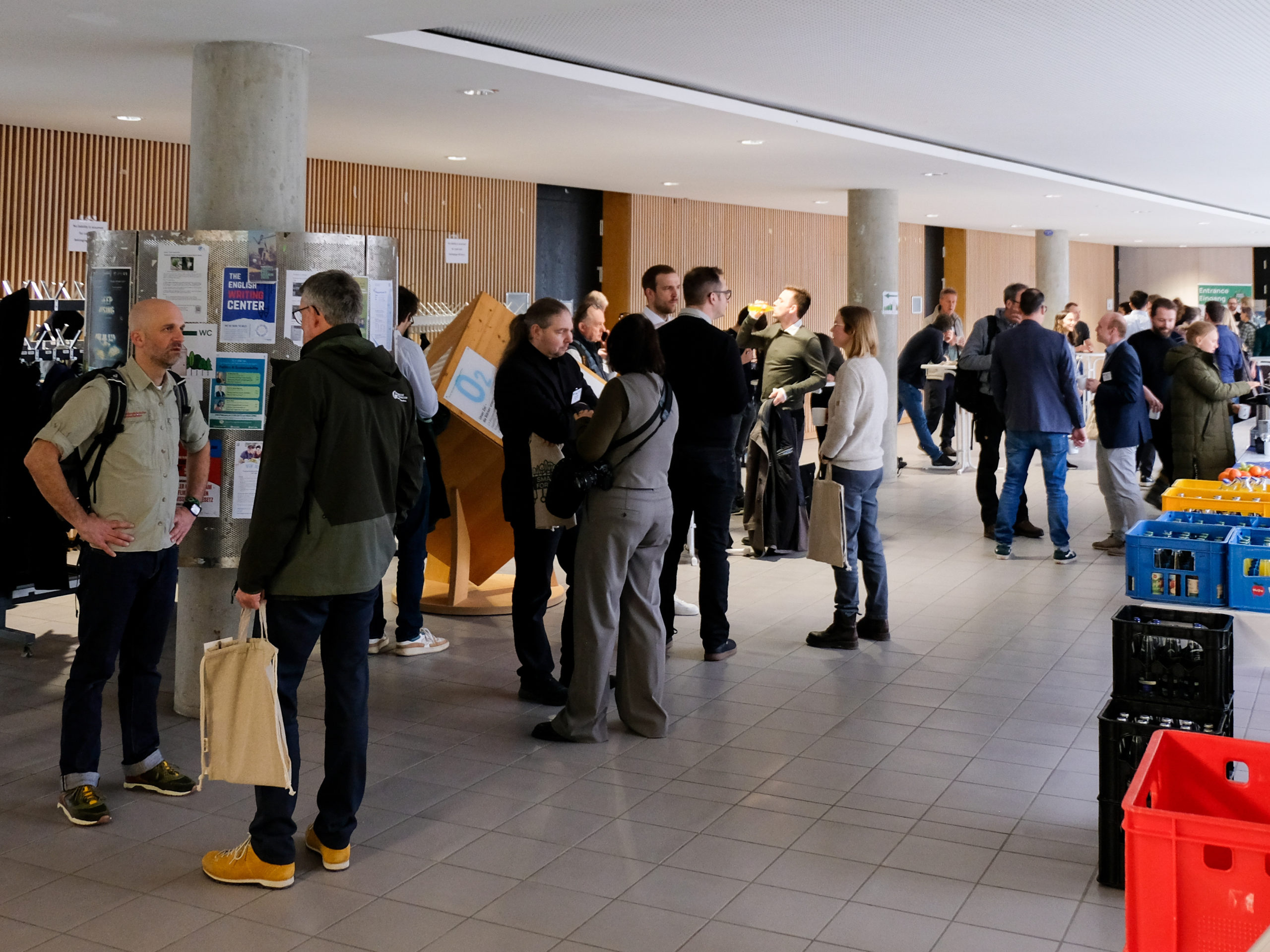 A crowd of people chats and has drinks in the foyer during SmartForest 2025. To the right, crates of drinks, as well as coffee urns, and fresh fruit are visible.