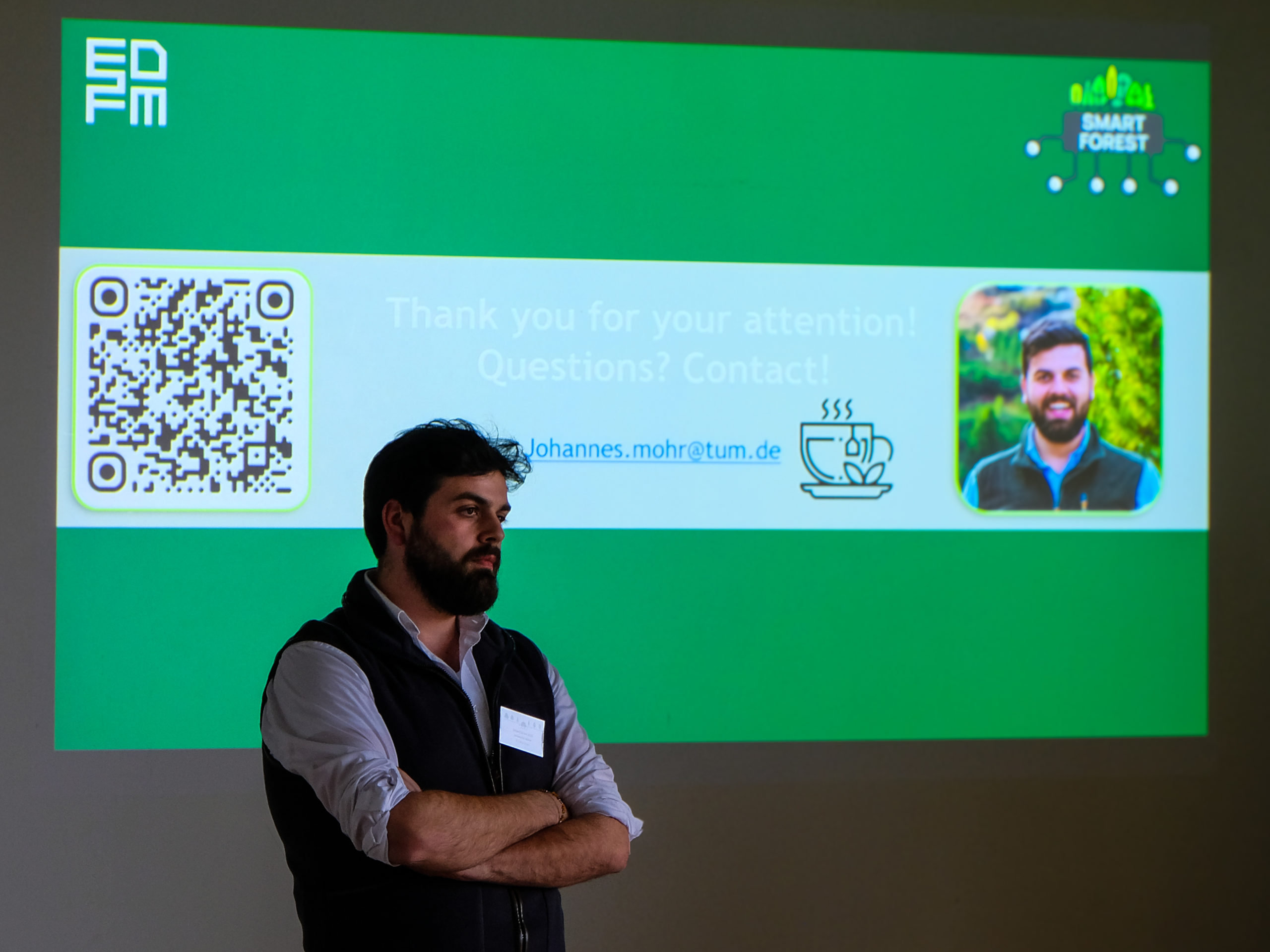 Johannes Mohr, a black-haired bearded man wearing a light blue-button up and black vest, stands with crossed arms as he listens to a question from the audience. Behind him, a "Thank you"-slide bears the branding of EDFM and SmartForest.