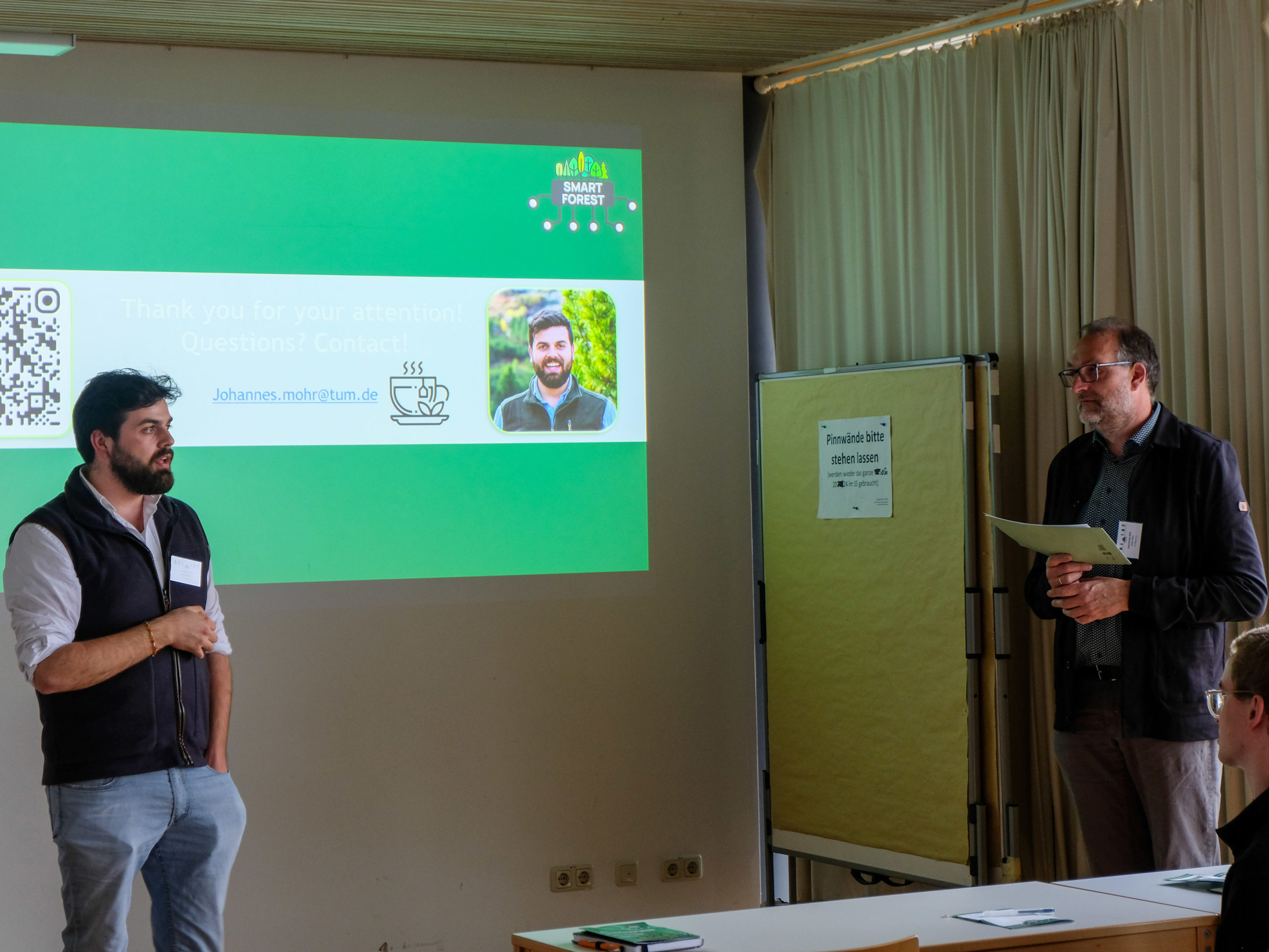 Johannes Mohr, a young bearded man with black hair, looks over at Dr. Peter Bieber, an older man with glasses and a greying beard, as he explains something in front of a SmartForest-branded slide with the text "Thank you for your attention! Questions? Contact?" along with his picture and contact information.