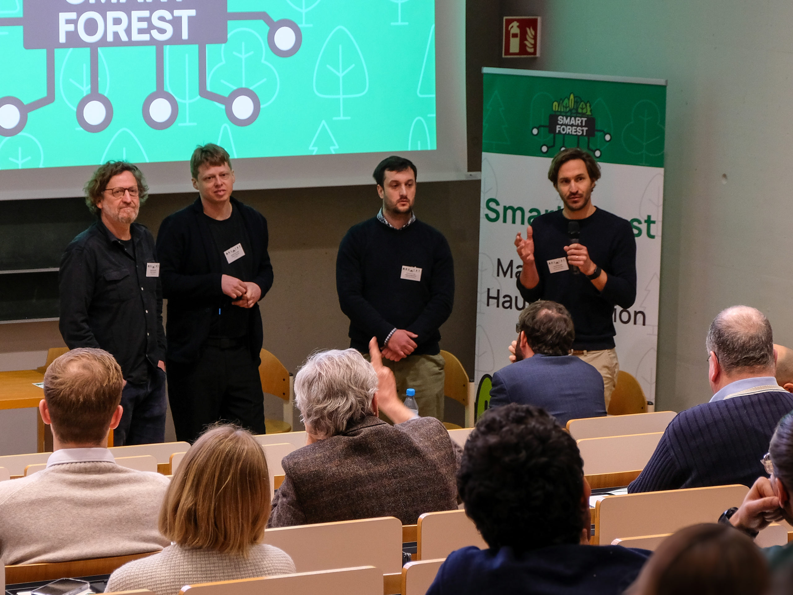 Dr. Werner Rammer, Dr. Fabian Fassnacht, Janusch Vajna-Jehlem and Dr. Felipe de Miguel Diez are at the front of the lecture hall. In the crowd, seen from the back, a grey-haired man in a brown jacket raises a finger.