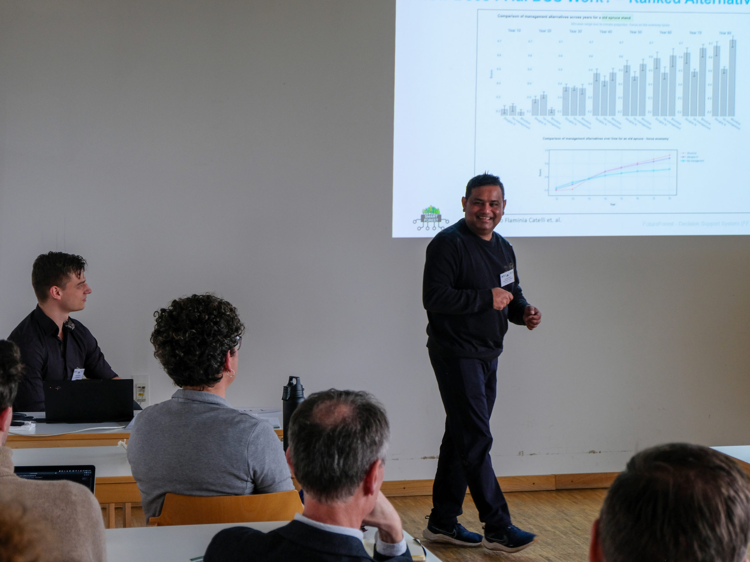 Dr. Somakanthan Somalingam, a dark-skinned man in a blue jumper, jeans, and sneakers, laughs in front of a slide full of data, as his curly-haired colleague Dr. Flaminia Catalli makes a remark from the front row.