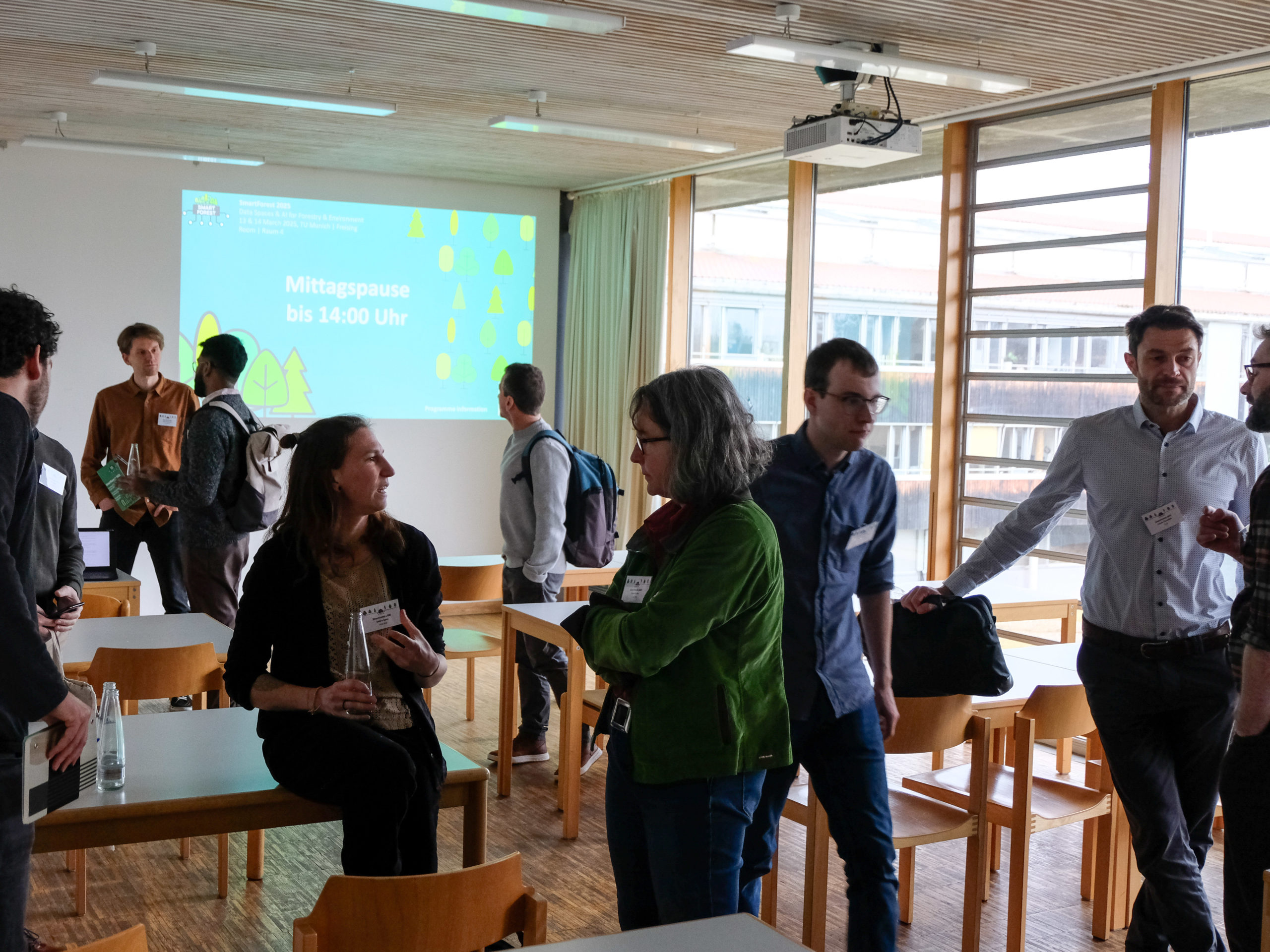 A small crowd of SmartForest 2025 attendees is gathered in a classroom with wooden tables and chairs. One wall is nearly entirely comprised of windows. They are speaking in small groups. In the background is a projected green slide with SmartForest-branding and the text "Mittagspause bis 14:00 Uhr"