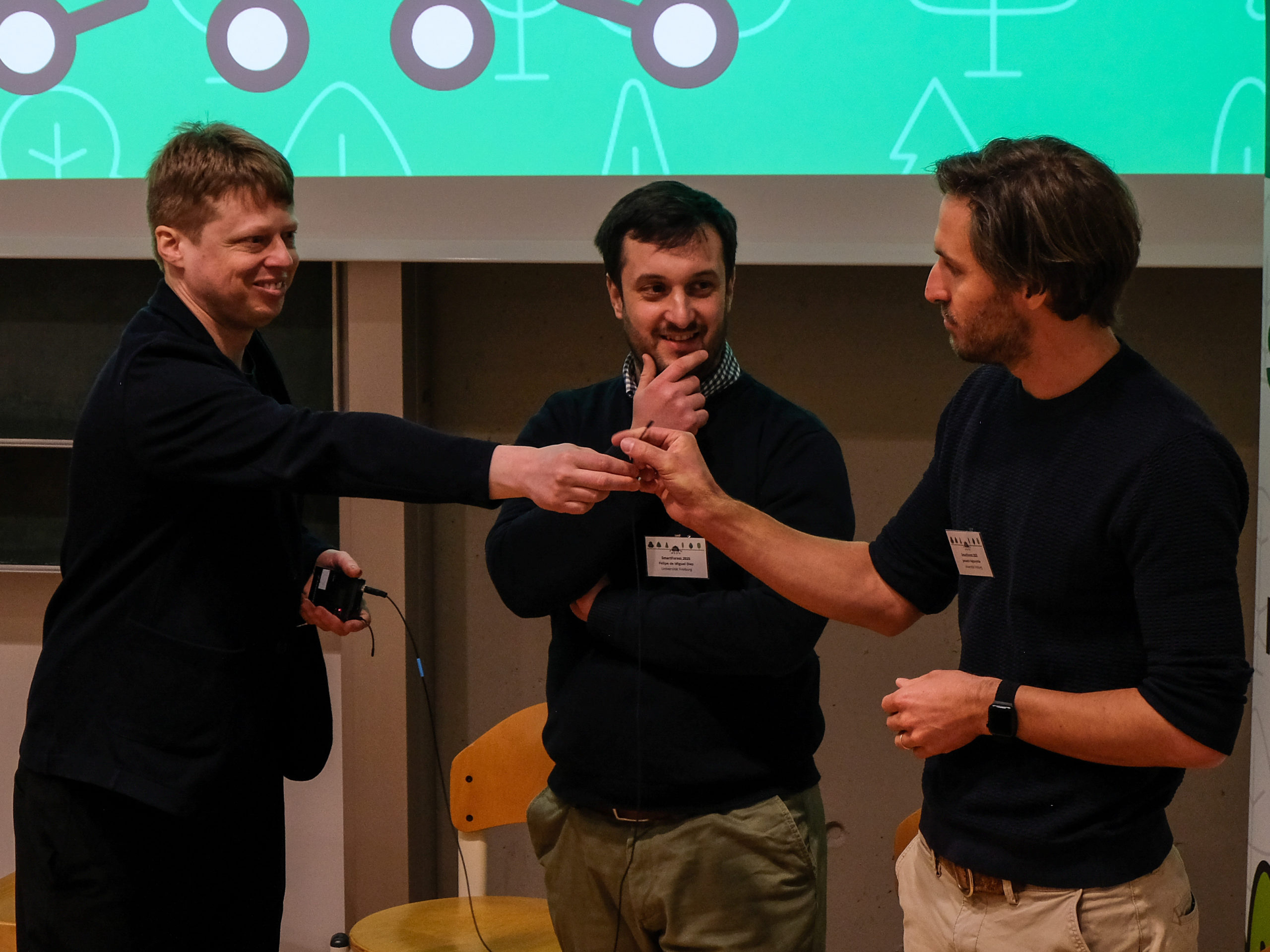 Prof. Dr. Fabian Fassnacht, a smiling blond man, passes a microphone to Janusch Vajna-Jehle, a bearded man with wavy dark hair, as Dr. Felipe de Miguel Diaz looks on.