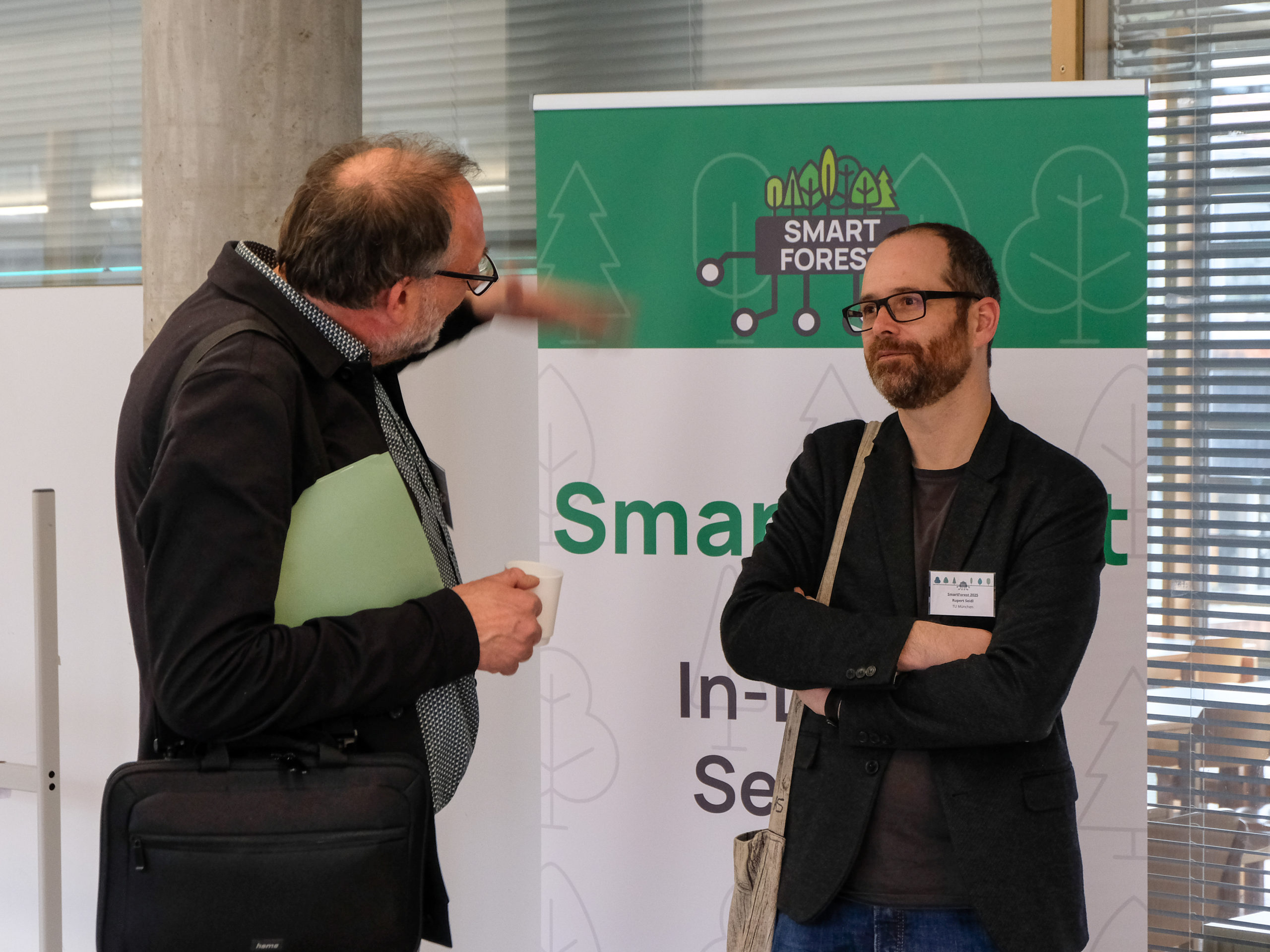 Dr. Rupert Seidl, a bespectacled man with short dark hair and beard, stands with his arms crossed in front of a SmartForest banner. To the left, another man in glasses, Dr. Peter Biber, is seen from the back, speaking to Prof Seidl. He is holding a mug in one hand whilst gesturing with the other.