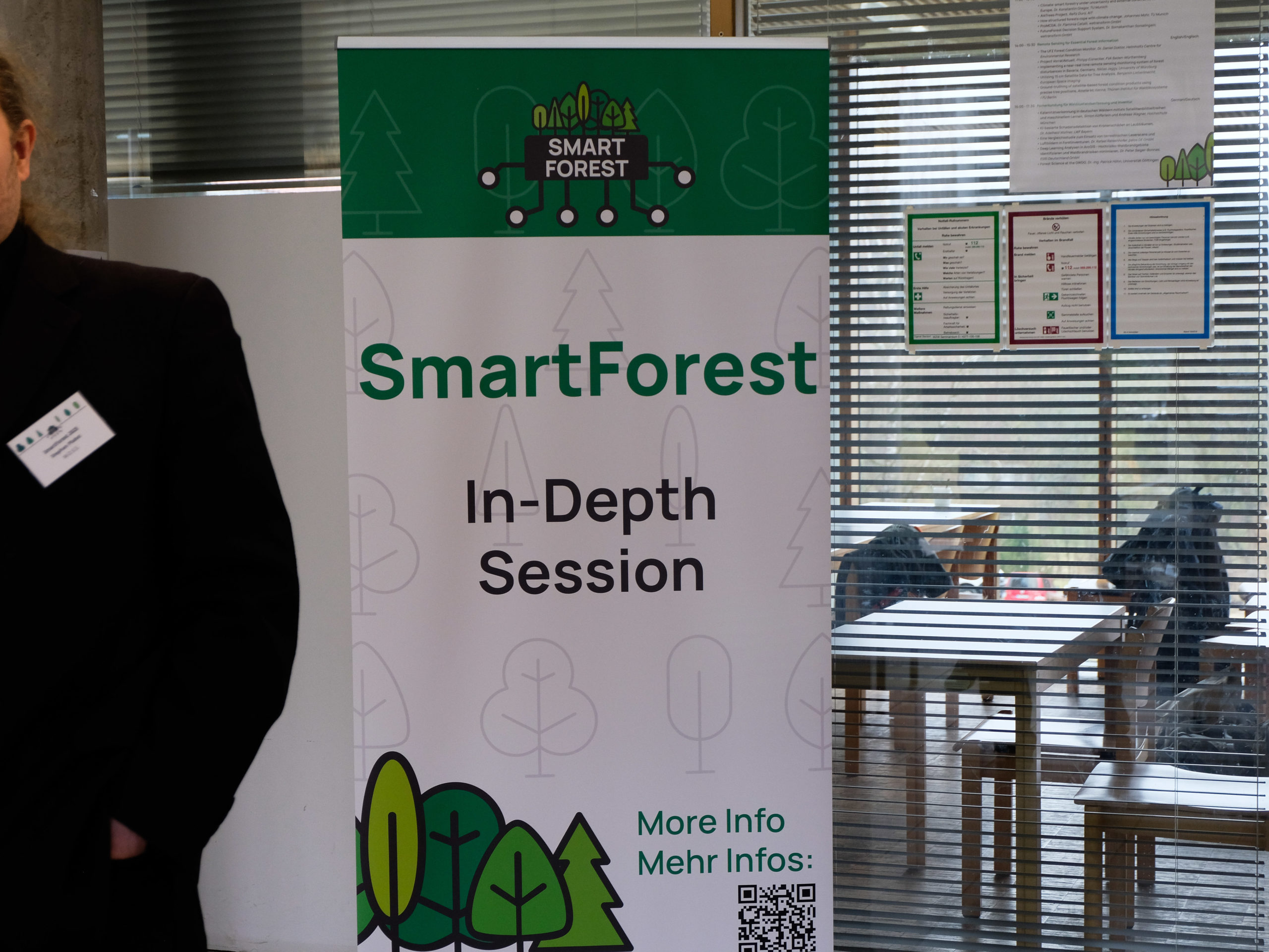A SmartForest-branded Roll-Up poster bears the text "In-Depth Session". To its right is a window with a view of a classroom with wooden tables and chairs and a large window to the outside. Safety instructions and a timetable have been taped to the internal window, but do not block it significantly. Coats and bags are visible on the tables and chairs. To the left is the shoulder of an attendee, with their SmartForest nametag just visible, but not legible.
