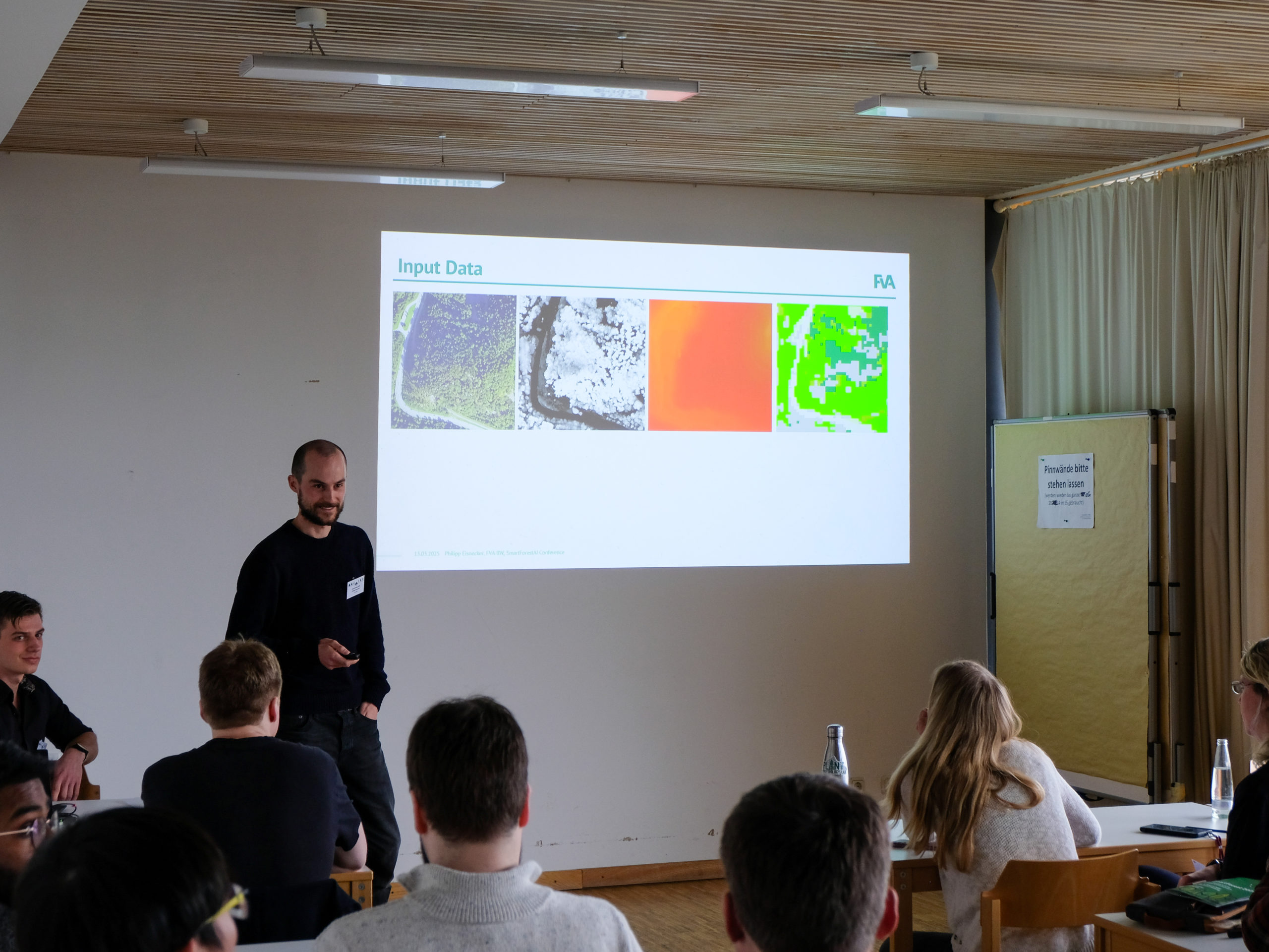 Philipp Eisnecker, a dark-haired man with a buzzcut and short beard, smiles as a blonde woman asks him questions from a classroom seat. Behind him is a slide with four pictures of the same aerial view of trees, each with different colours, titled "Input Data".