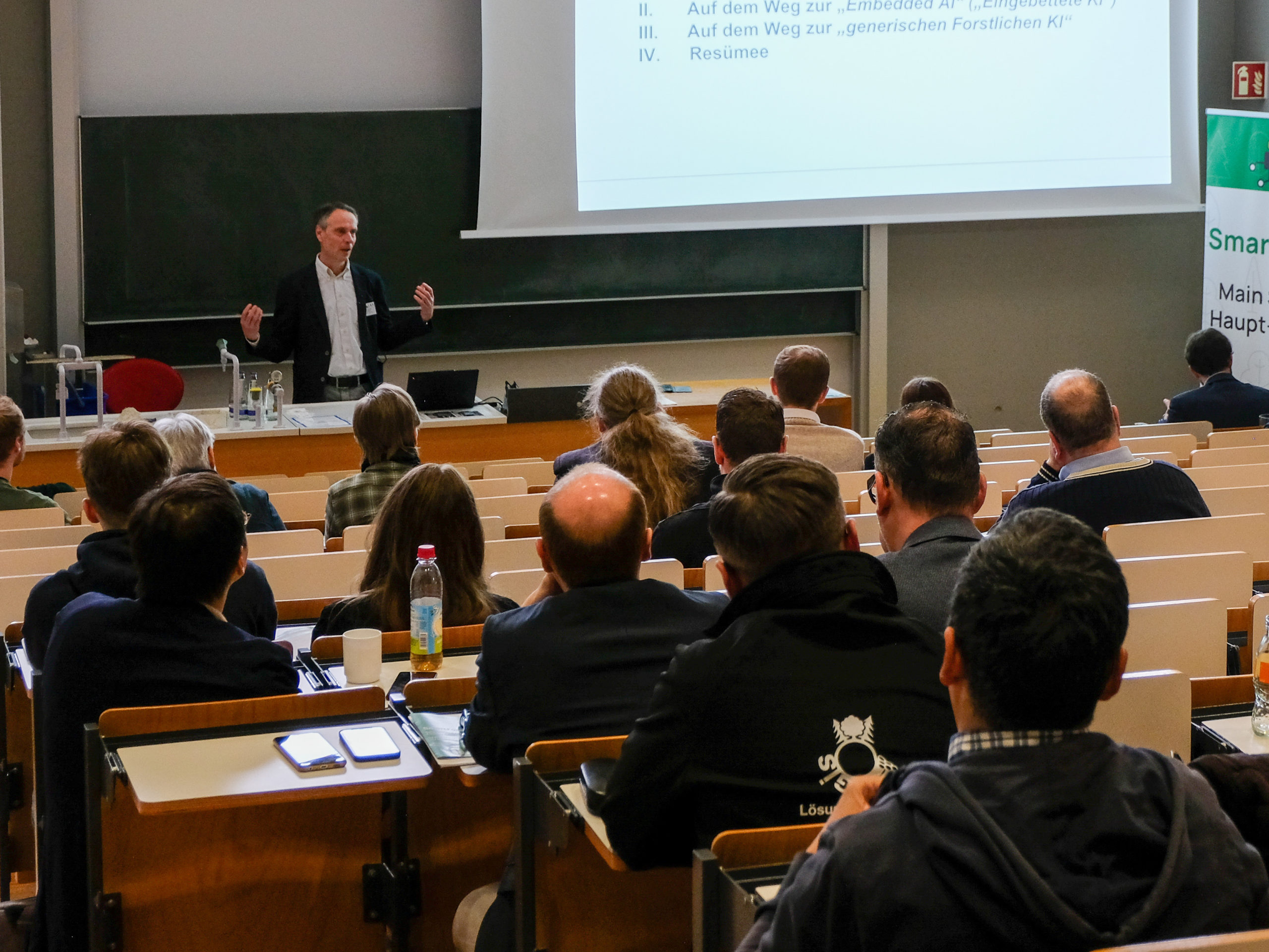 Prof. Dr. Martin Ziesak, sporting a light button-up and dark jacket, is seen lecturing to an attentive audience. Only part of the current slide is visible, which reads "II. Auf dem Weg zur Embedded AI (Eingebettete KI), III. Auf dem Weg zur generischen Forstlichen KI, IV. Resümee"