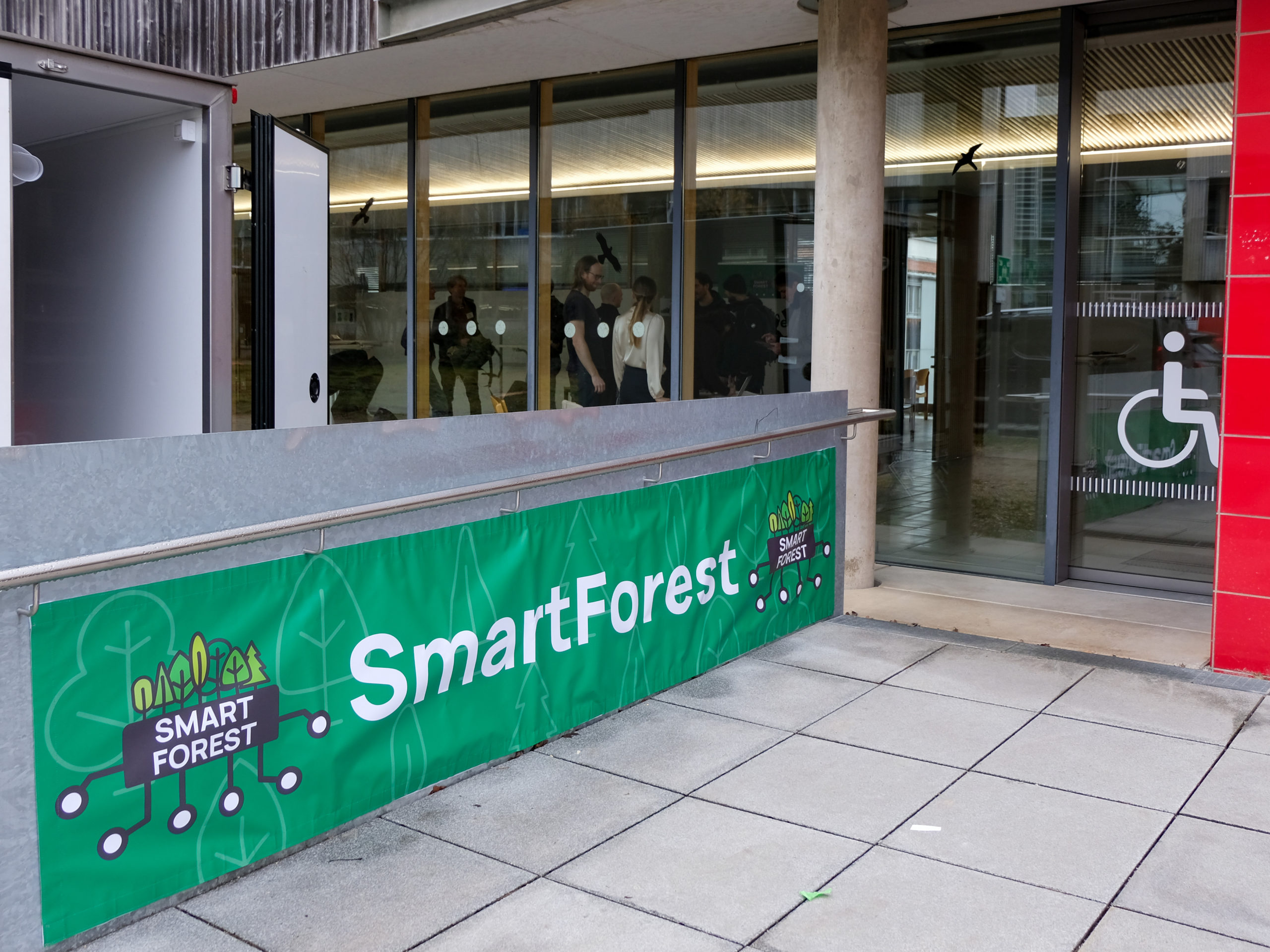 The accessibility-friendly entrance to SmartForest 2025. In the foreground, below a handrail, is a large green banner with the SmartForest-branding. to the right is an automated sliding door with a pictogram of a person in a wheelchair. Through the building's windows, members of the SmartForest team can be seen.