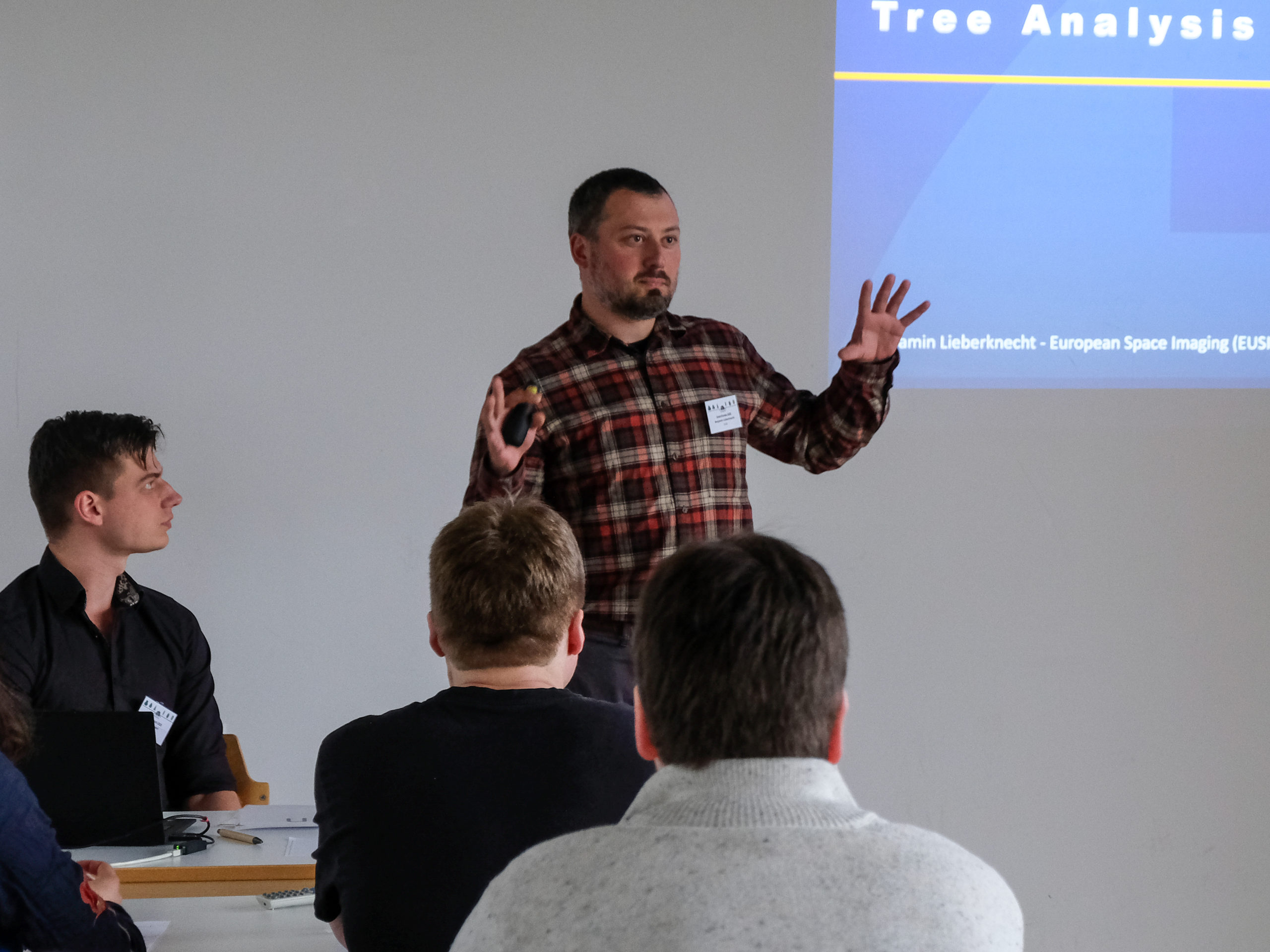 Benjamin Lieberknecht, a bearded man in a red checked button-up explains something to an interested classroom. Behind him, a partially visible projection shows the words "Tree Analysis" and "European Space Imaging".