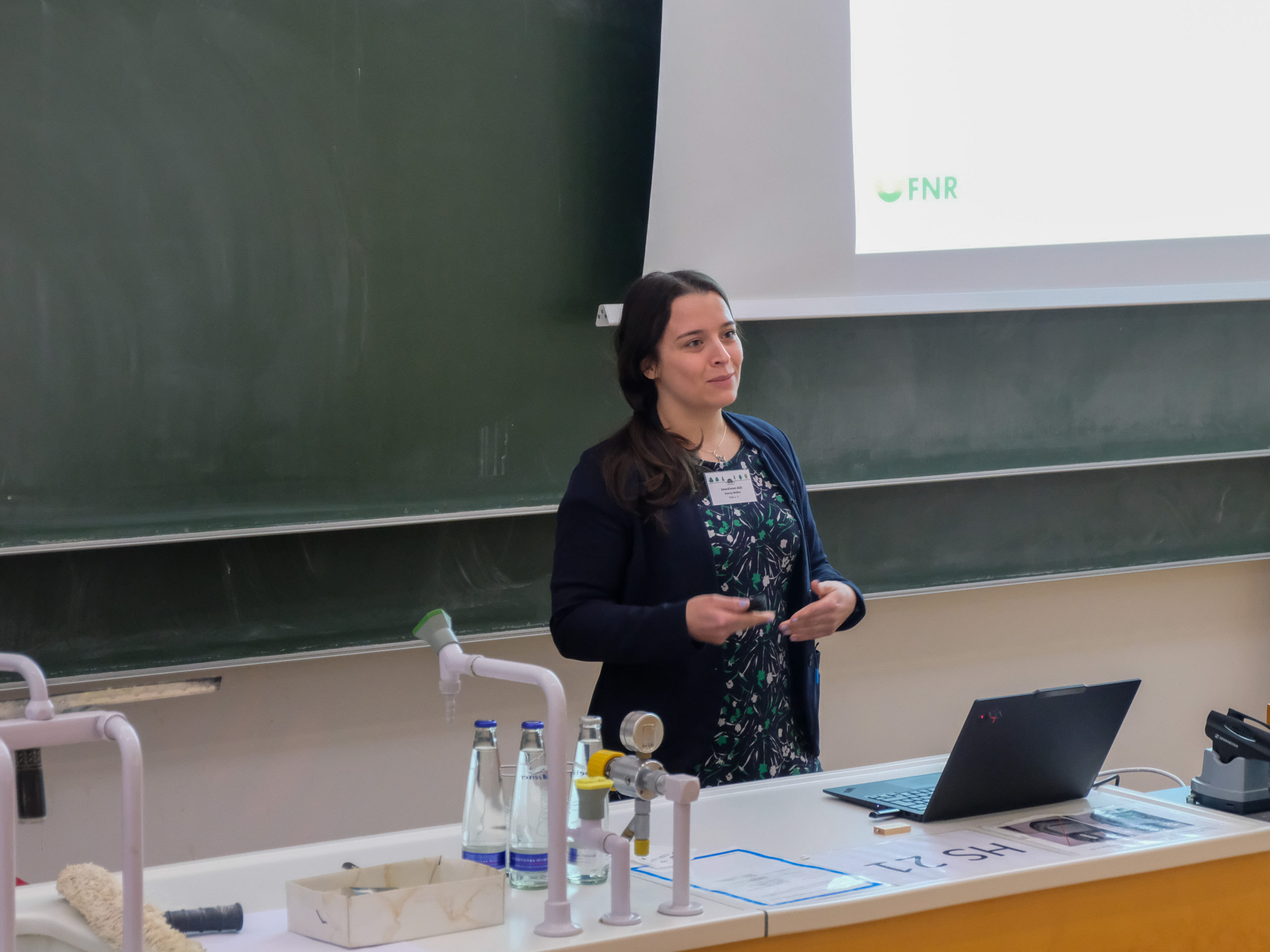 Nancy Müller, a dark-haired woman in a dark dress with abstract plant-motif and a blue cardigan explains something in front of a blackboard. On a projection behind her, the "FNR"-logo of Fachagentur Nachwachsende Rohstoffe is visible.