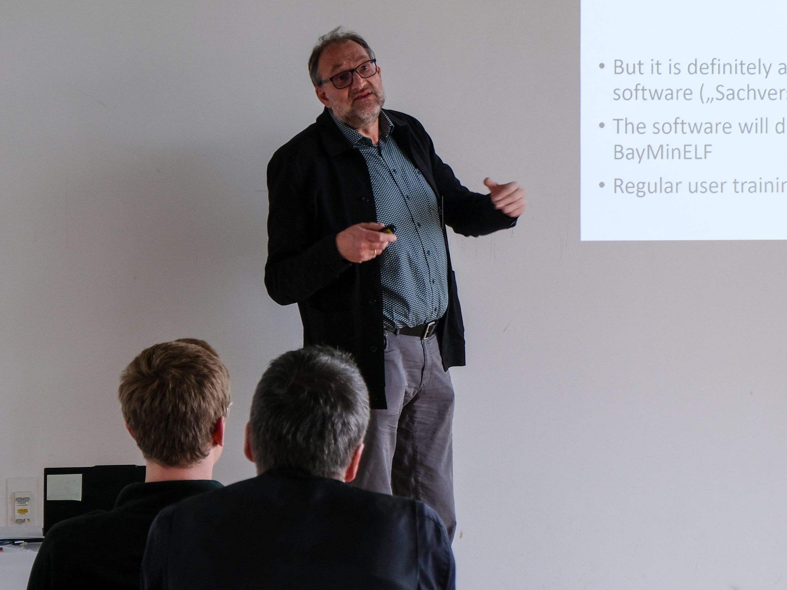 A bespectacled man with a short, greying beard explains something to people in a classroom.