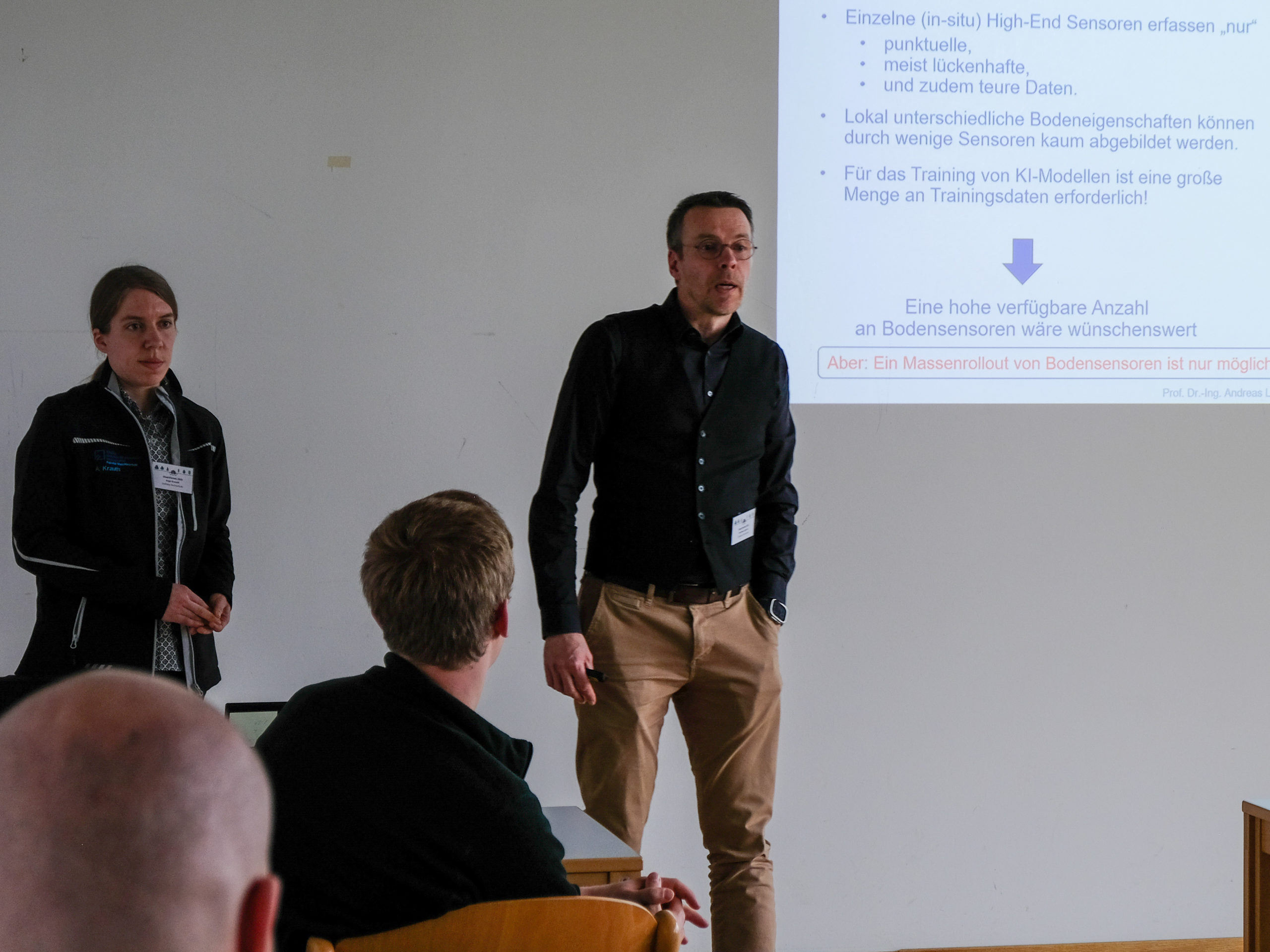 Prof. Dr.-Ing. Andreas Ligocki, a tall bespectacled man, and Anja Krauth, a woman with a dark ponytail, stand in front of a classroom, with onlookers seen from the back. Prof. Ligocki appears to be in the middle of answering a question. Behind him, a projected slide is partially visible and reads: "Einzelne (in-situ) High-End Sensoren erfassen "nur" - punktuelle, - meist lückenhafte, - und zudem teure Daten. Lokal unterschiedliche Bodeneigenschaften können durch wenige Sensoren kaum abgebildet werden. Für das Training von KI-Modellen ist eine große Menge an trainingsdaten erforderlich!", a blue arrow points down at a further bit of blue text: "Eine hohe verfügbare Anzahl an Bodensensoren wäre wünschenswert", followed by a red: "Aber: Ein Massenrollout von Bodensensoren ist nur möglich", with the rest cut off.