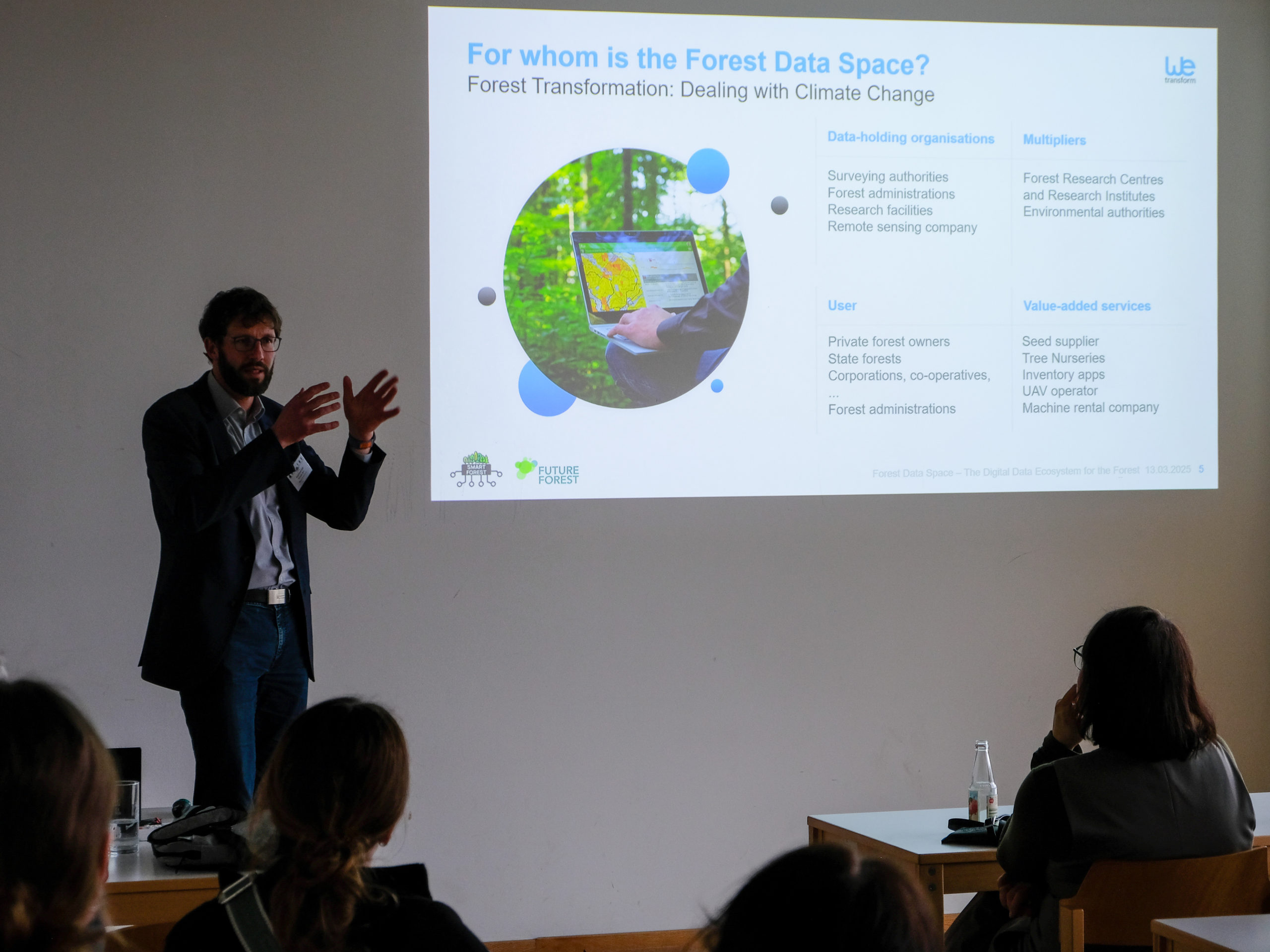 Thorsten Reitz, a tall man with a beard and glasses, gestures as he explains something to a crowd of fascinated onlookers. A projected PowerPoint slide beside him asks the question "For whom is the Forest Data Space?", with the subtitle "Forest Transformation: Dealing with Climate Change". It answers the question with four lists of Data-holding Organisations, Multipliers, User, and Value-added Services.