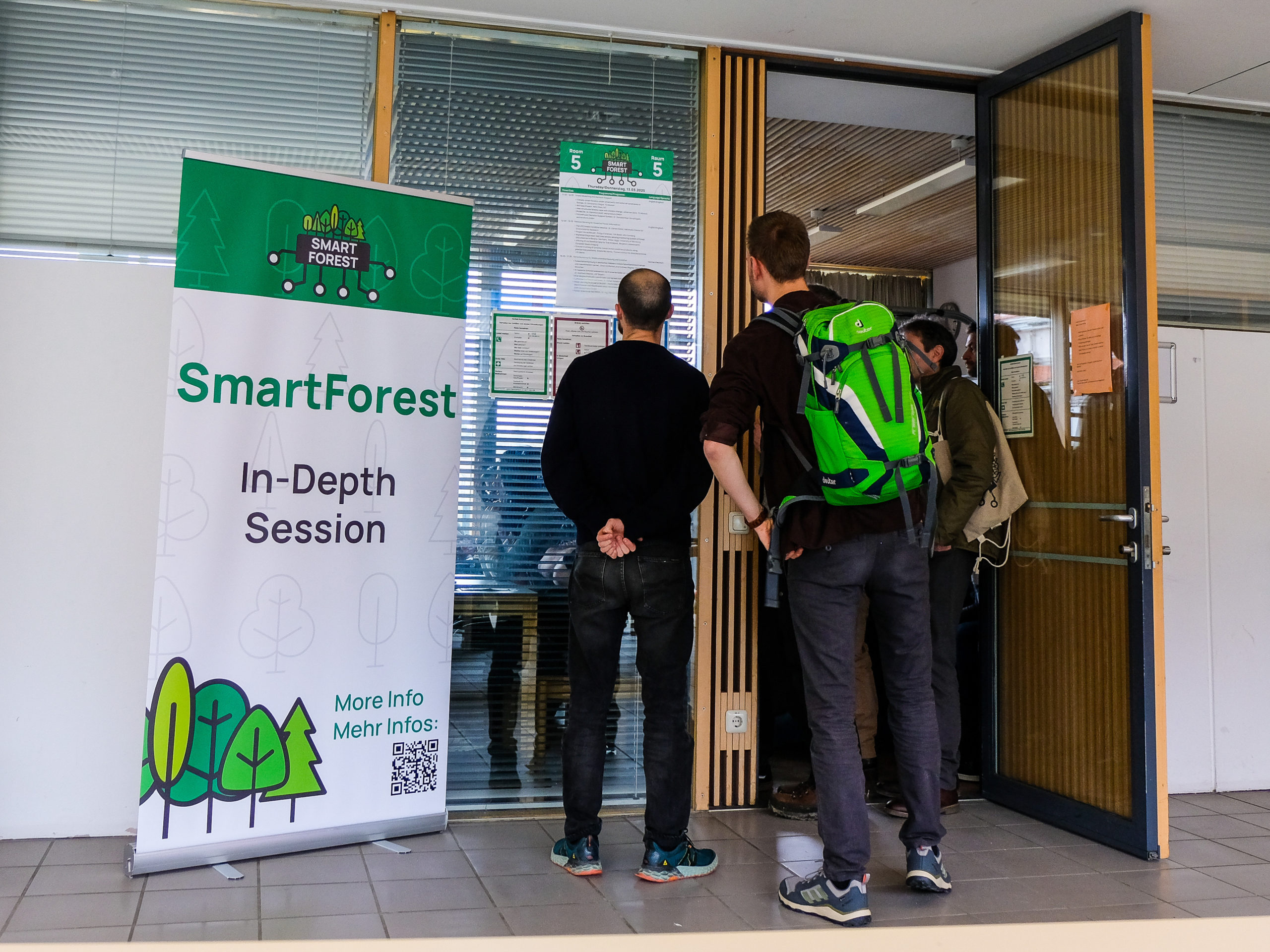 A small crowd of people is gathered around the door of Room 5, their backs to the camera as they inspect a poster bearing the room's schedule for the day. To their left is a SmartForest banner bearing the words "In-Depth Session".