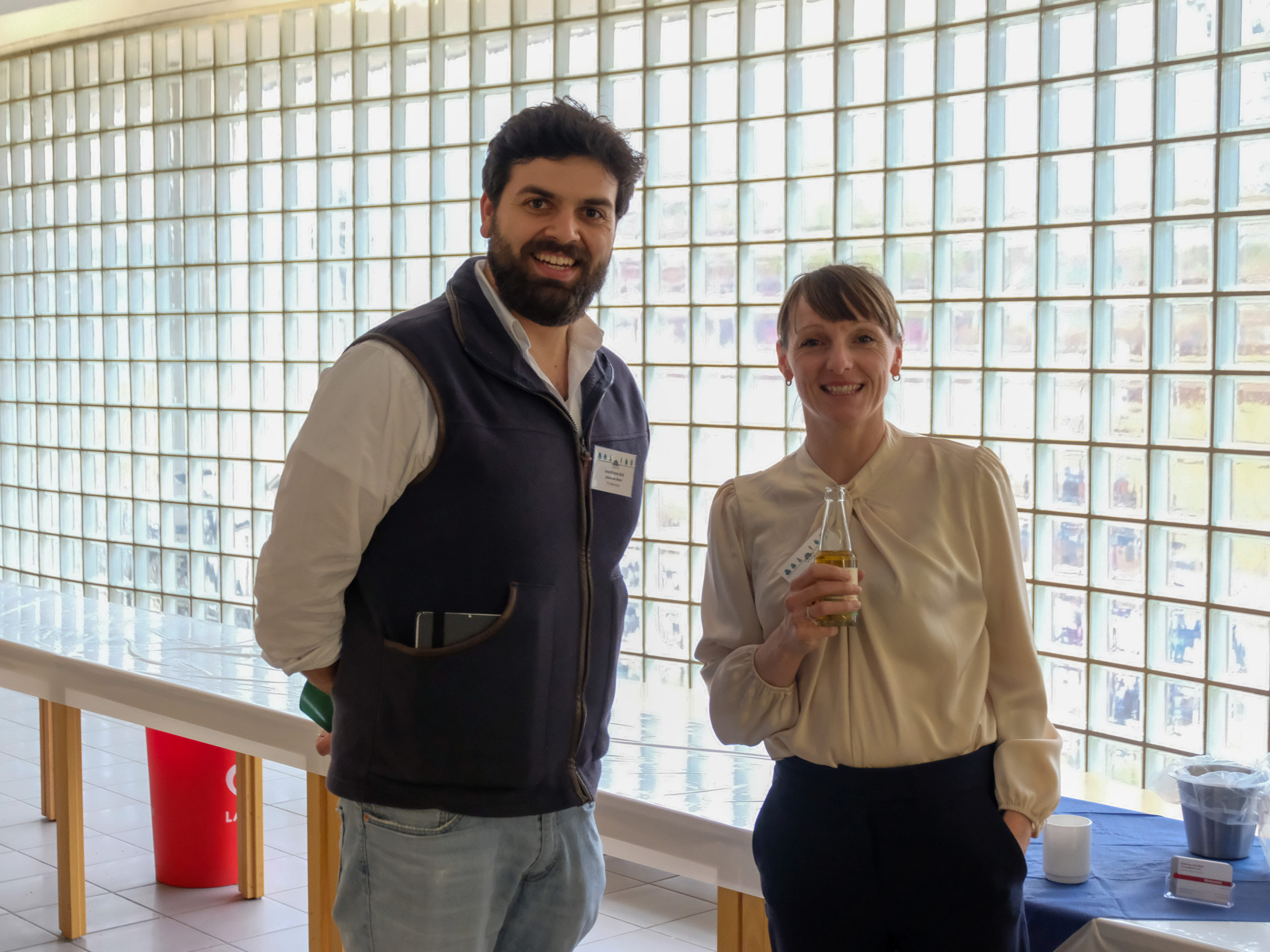 Two members of the SmartForest organisation team smile at the camera in the conference's foyer, a glass wall behind them. To the left, Johannes Mohr, a tall bearded man, and to the right, Franziska Hochenegger, a blonde woman with a ponytail holding a bottle of juice.