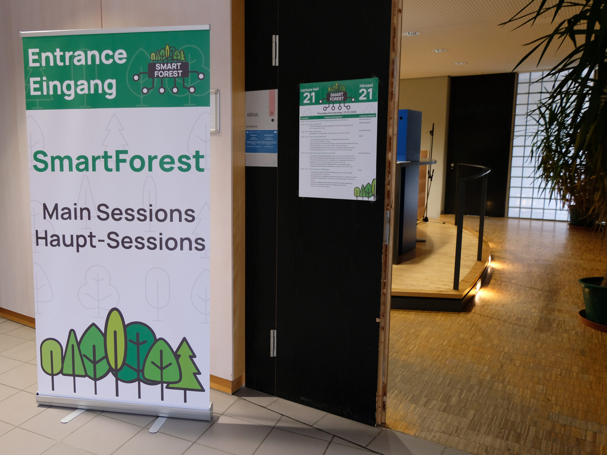 The doorway to Lecture Hall 21 at SmartForest 2025. Plants can be seen through the open door, while on the panel next to it is a poster showing the time schedule for that room on Thursday, March 13th. To the left is a large, SmartForest-branded stand-up poster that bears the text "Entrance Eingang. SmartForest. Main Sessions Haupt-Sessions"