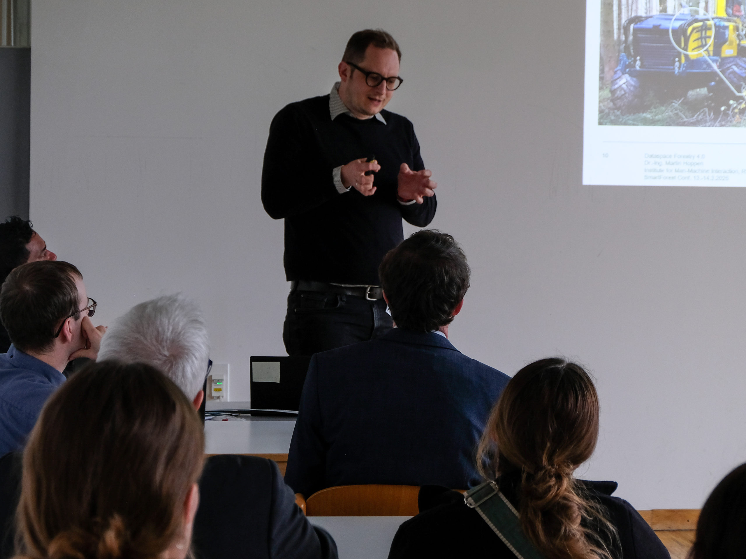 Dr. Martin Hoppen, a dark-haired man sporting heavy-rimmed glasses and a dark jumper over a light button-down gestures in front of his chest, palms cupped towards each other as if grasping something, A classroom of interested people look on. A projected slide showing forestry equipment is just visible to the right of the picture.
