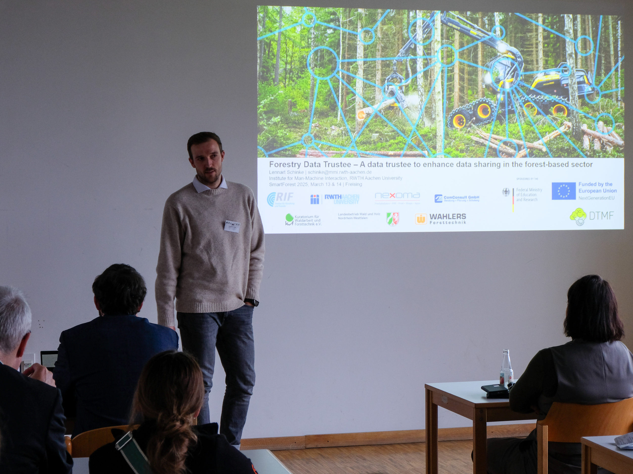 Lennart Schinke, a dark-haired man with a stubbly beard and light-coloured jumper, stands in front of a classroom, listening to someone as they ask a question. Beside him is a slide showing forestry equipment and many logos. It is titled "Forestry Data Trustee - A data trustee to enhance data sharing in the forest-based sector."