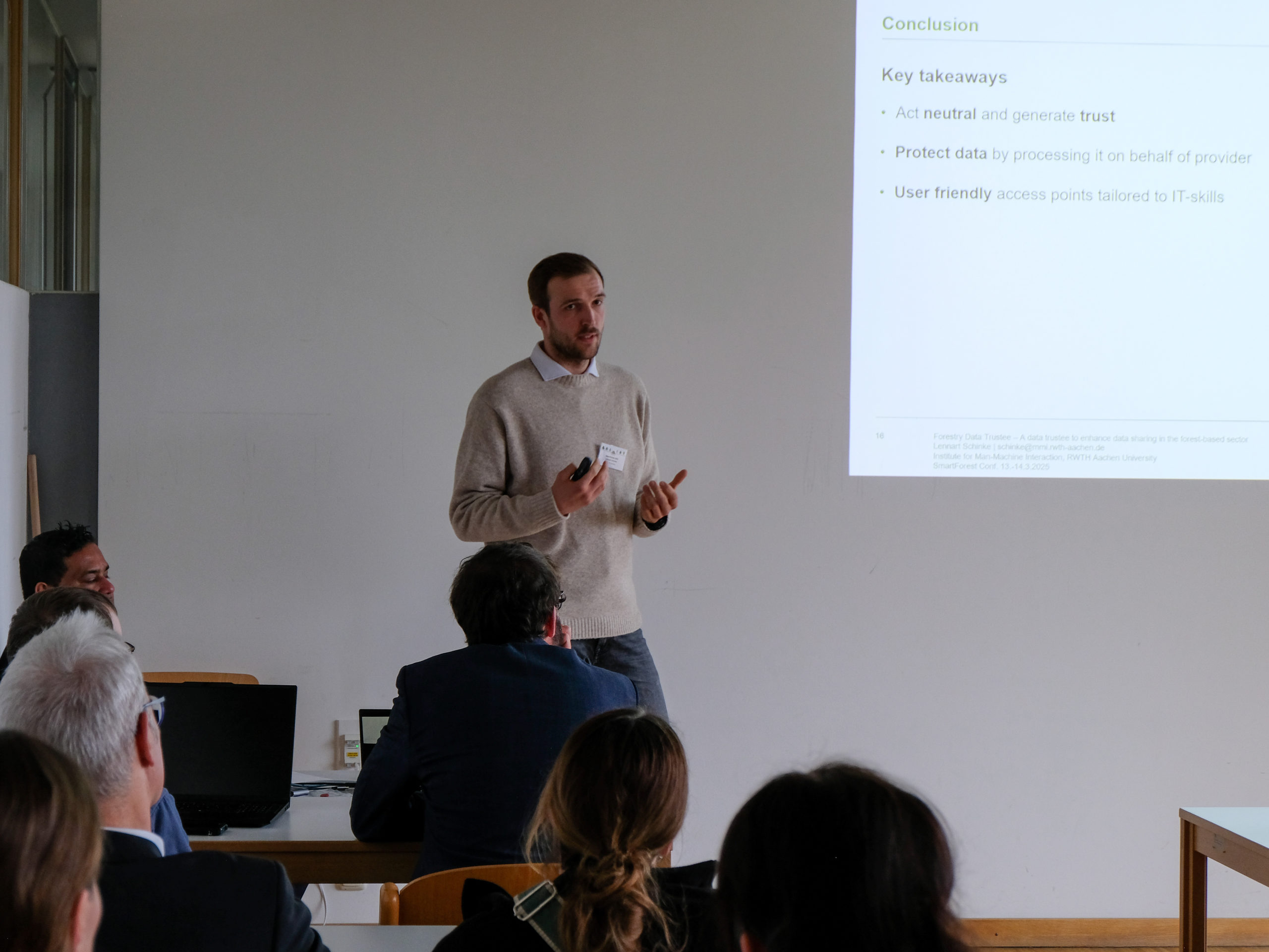 Lennart Schinke, a dark-haired man with a stubbly beard and light-coloured jumper, stands at the front of a classroom, gesturing as he explains something to a crowd of interested onlookers. A projected slide next to him reads "Conclusion. Key takeaways. Act neutral and generate trust. Protect data by processing it on behalf of provider. User friendly access points tailored to IT-skills"