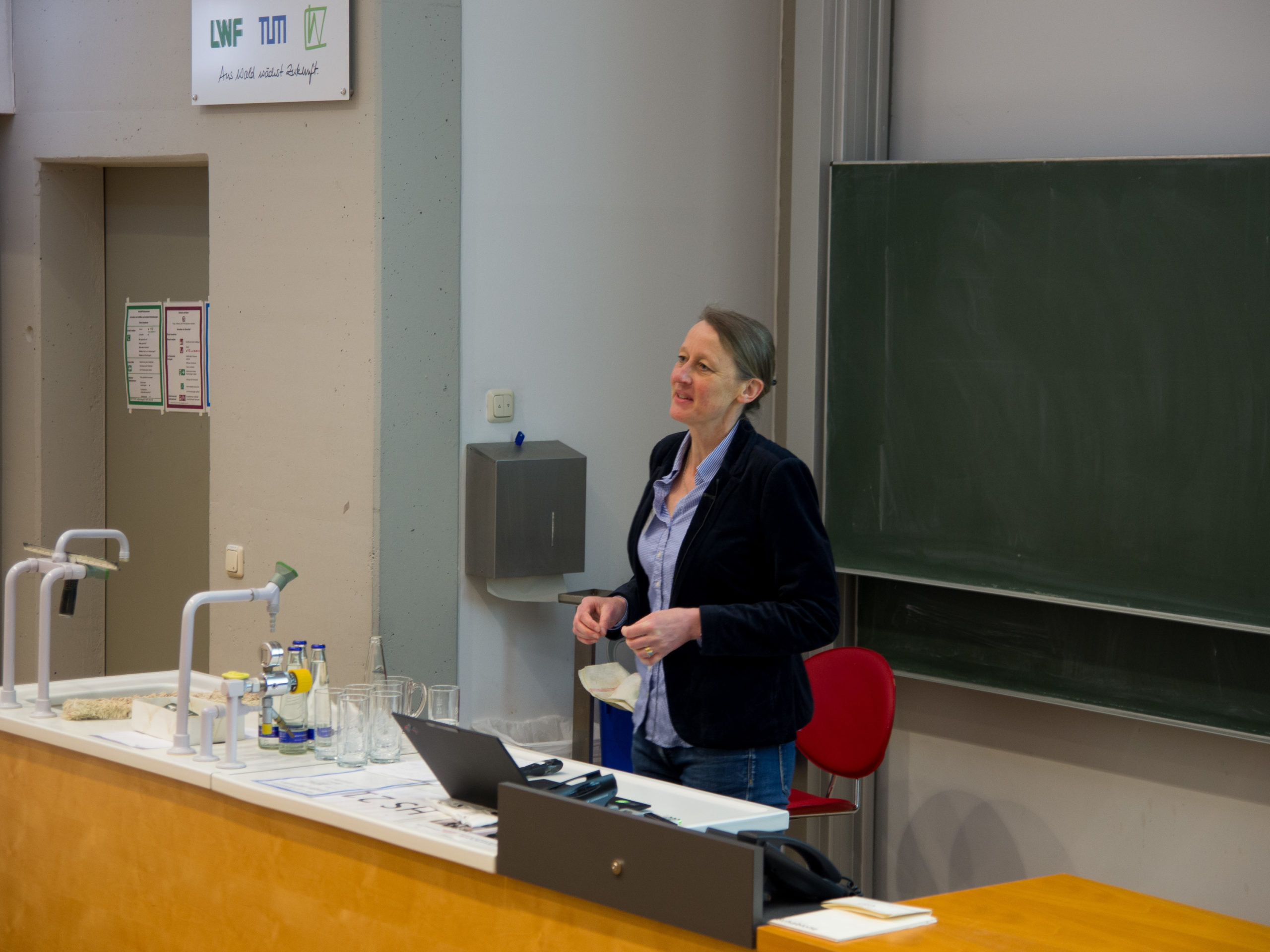 Dr. Ruth Dirsch, a woman in a light blue blouse, darker blue blazer, and jeans, addresses the SmartForest crowd in front of a blackboard.