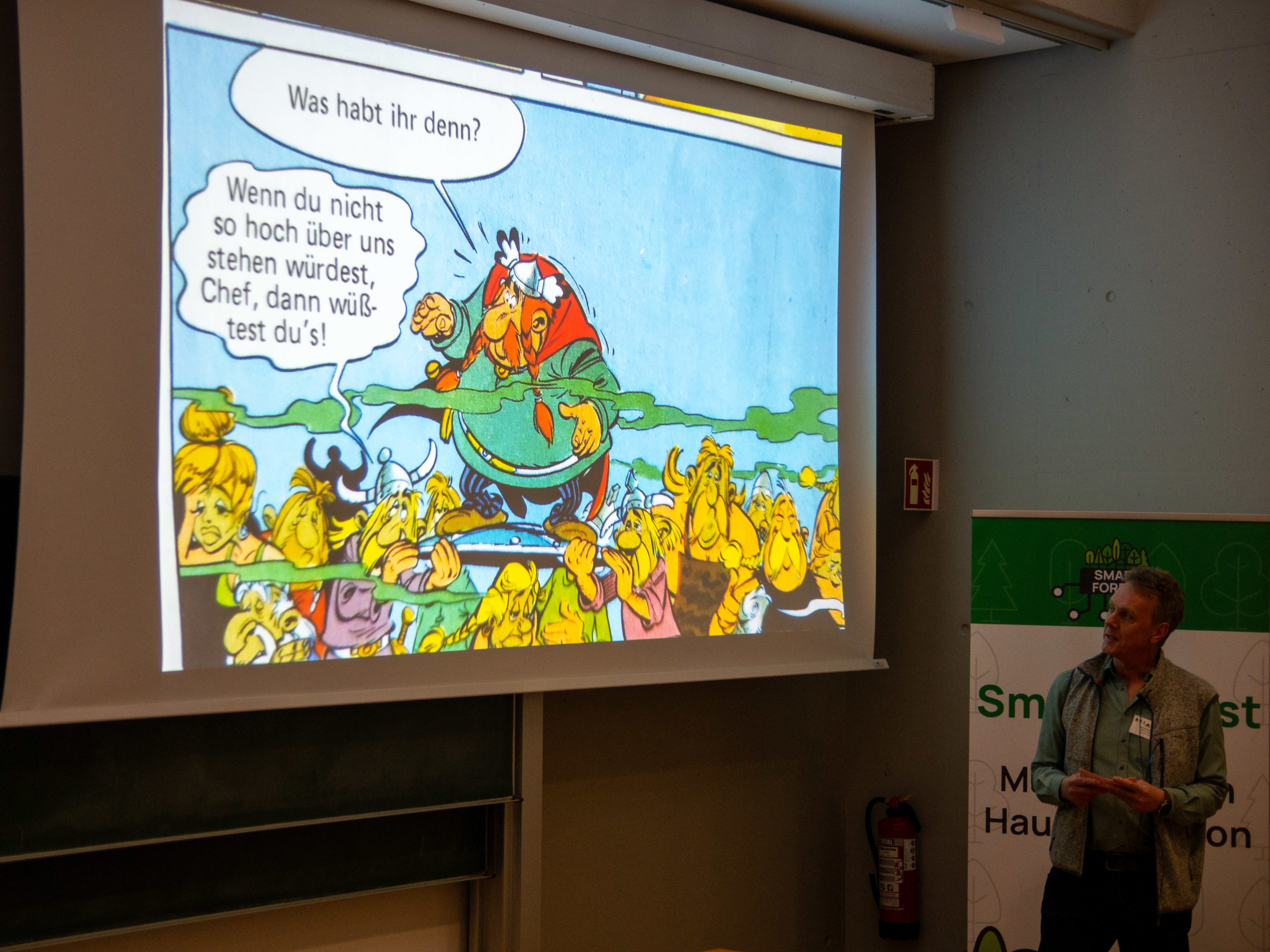 A man stands in front of a SmartForest poster, lecturing. To the left is a projection of an Asterix comic panel in German. Vitalstatistix, the red-headed Gaulish chief, is standing up, carried atop a shield. Just below his chest-level, green "stinky" vapour lines are visible. He asks the crowd of green-faced villagers below him "Was habt ihr denn?", to which one of his carriers replies "Wenn du nicht so hoch über uns stehen würdest, Chef, dann wüßtest du's!"