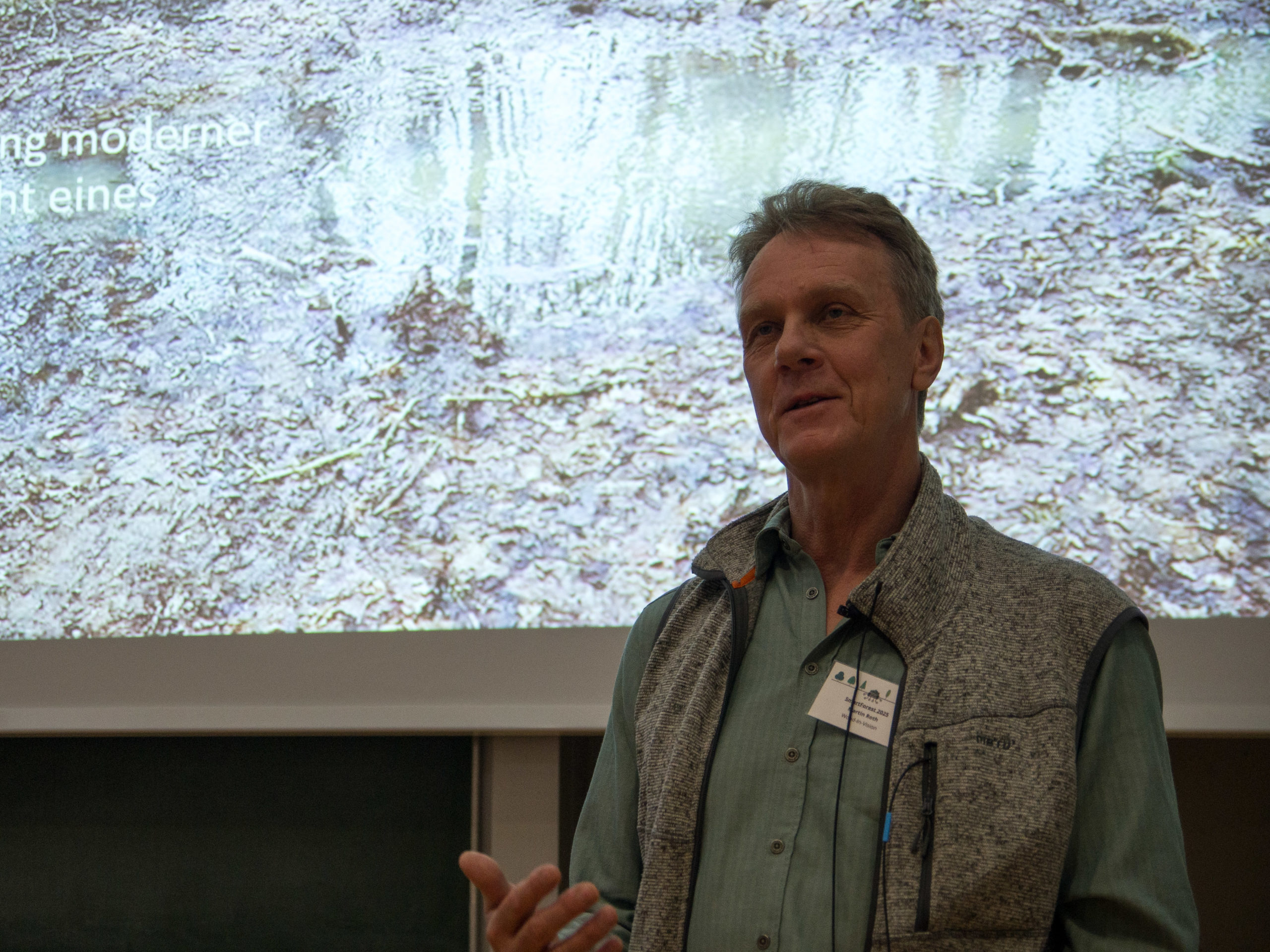 A man in a green button-up and grey vest stands in front of a projection of a forest floor. He appears to be calmly explaining something. His SmartForest 2025 name tag proclaims him to be Martin Roth of Wood-in-Vision.