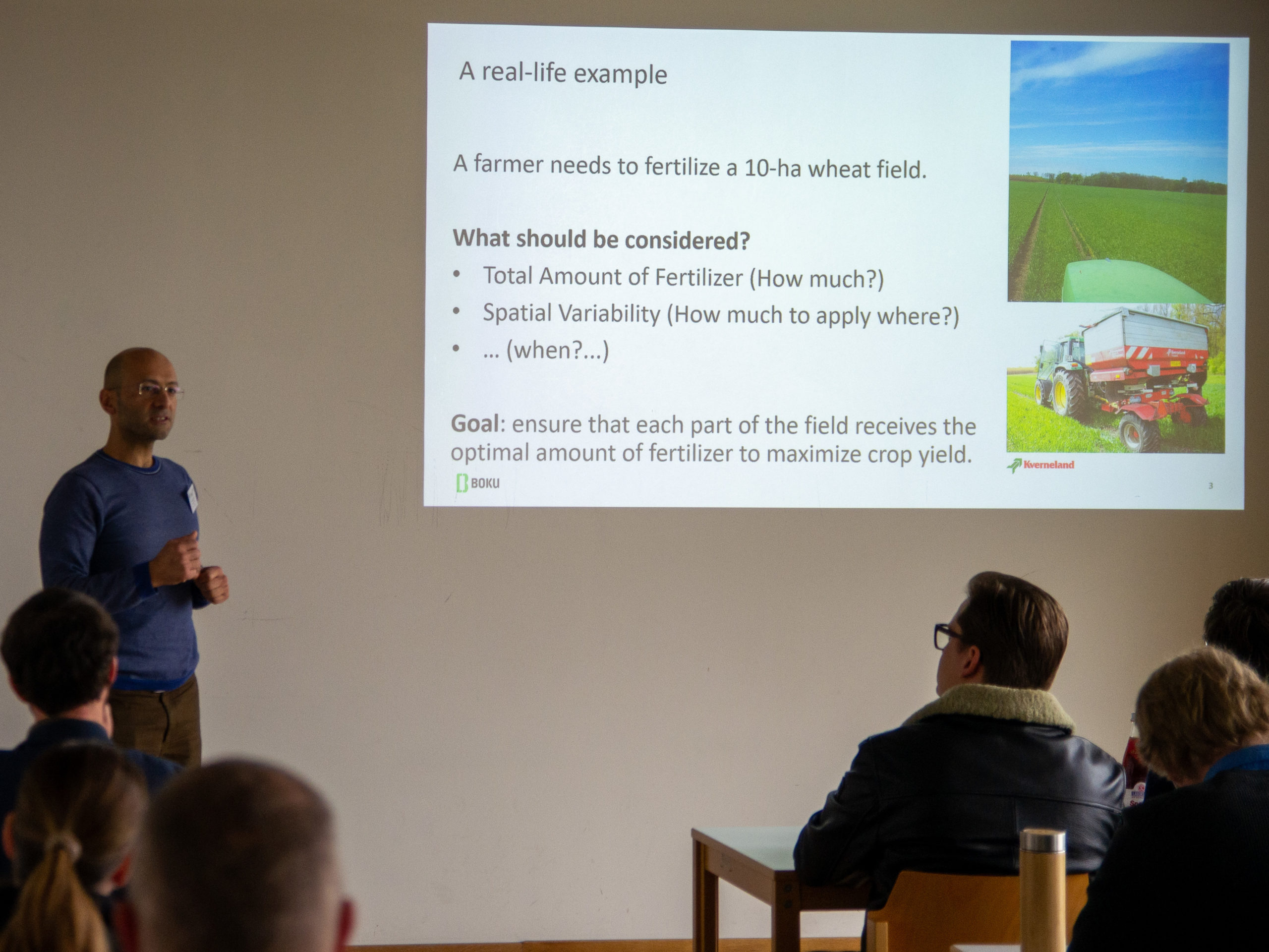 A bald man in glasses addresses a crowd of people in a classroom. Next to him is a projected slide, sporting the logos of BOKU and Kverneland, as well as two pictures of tractors and some text. The text reads: "A real-life example. A farmer needs to fertilize a 10-ha wheat field. What should be considered? - Total Amount of Fertilizer (How much?), - Spatial Variability (How much to apply where?), - ... (when?...) Goal: ensure that each part of the field receives the optimal amount of fertilizer to maximize crop yield."