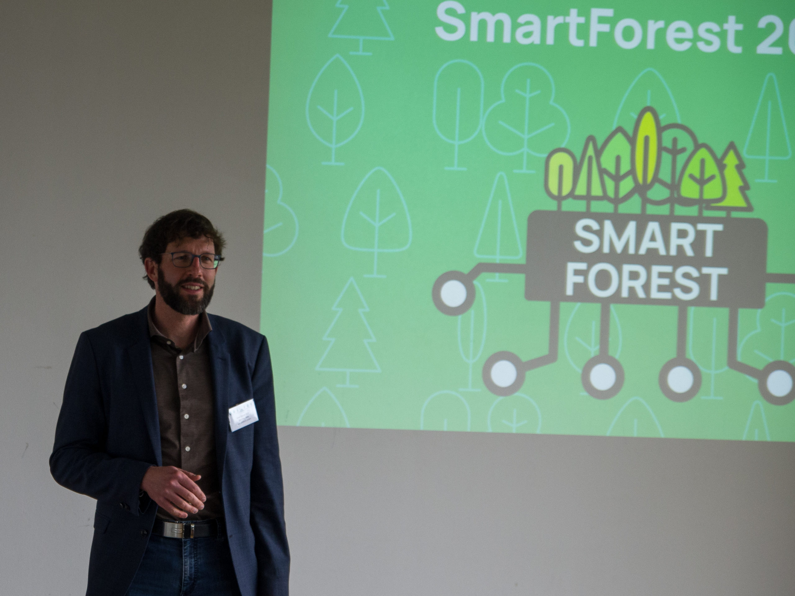 Thorsten Reitz, a tall, dark-haired. bearded man with glasses, wearing a dark button-down and blue jacket, stands next to a large projection of the SmartForest logo on a green background with stylised white trees.