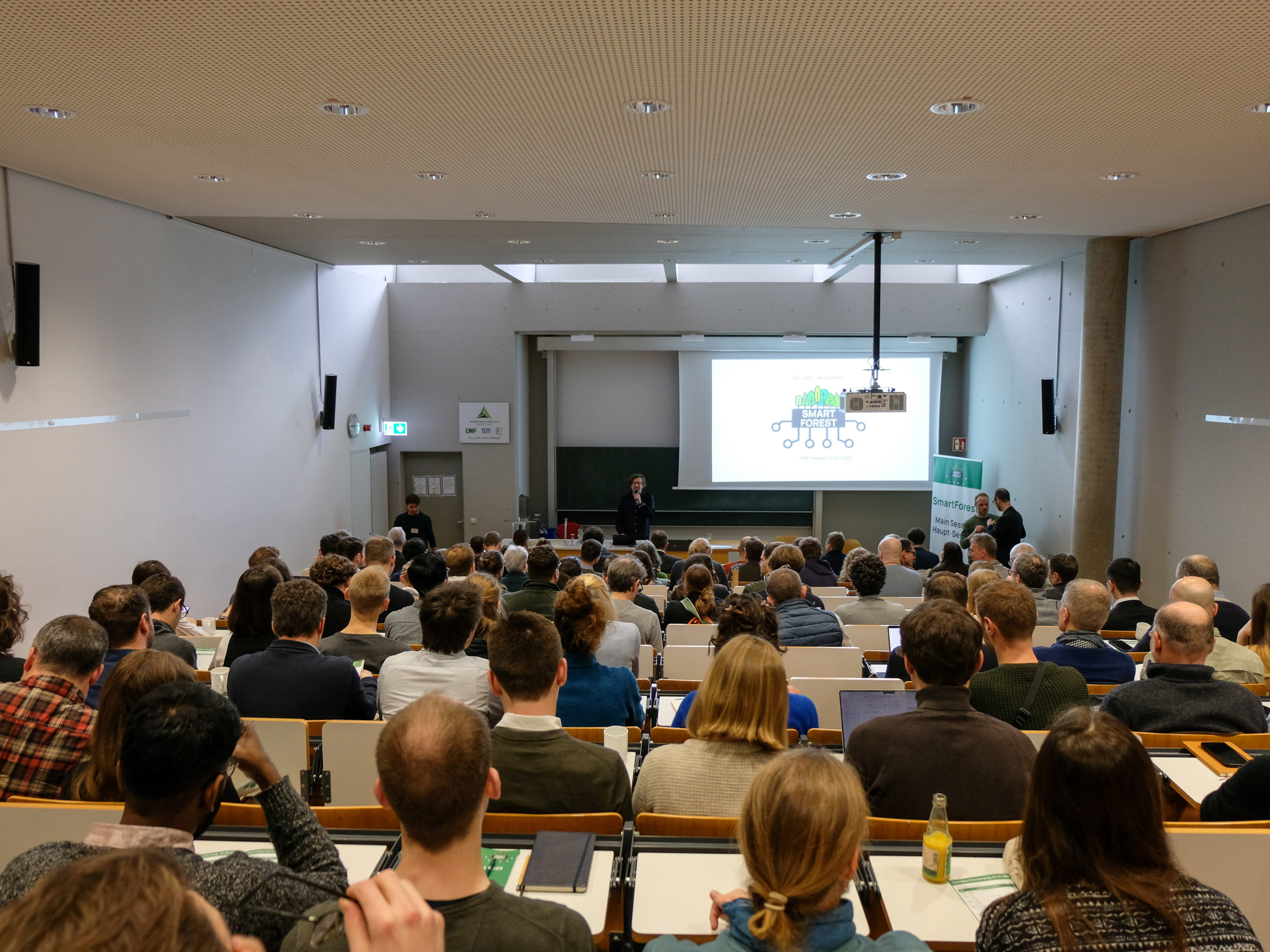 Photograph taken from the back of the full lecture hall during the opening of SmartForest 2025. The crowd listens attentively to Dr. Werner Rammer while Prof. Dr. Rupert Seidl's microphone is being hooked up on the right.