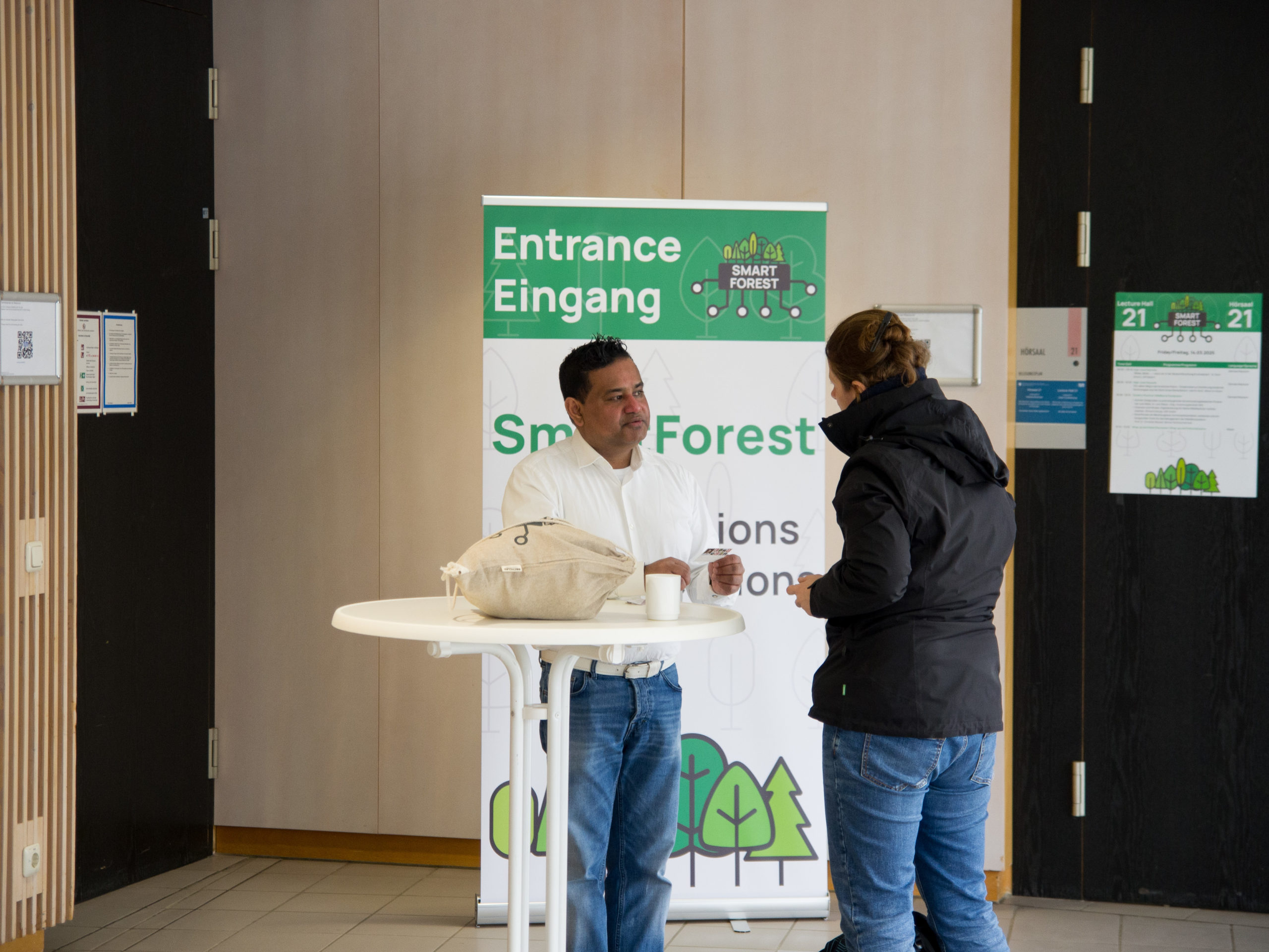 Two SmartForest 2025 participants have a discussion whilst standing by a white table in front of the conference's welcome sign.