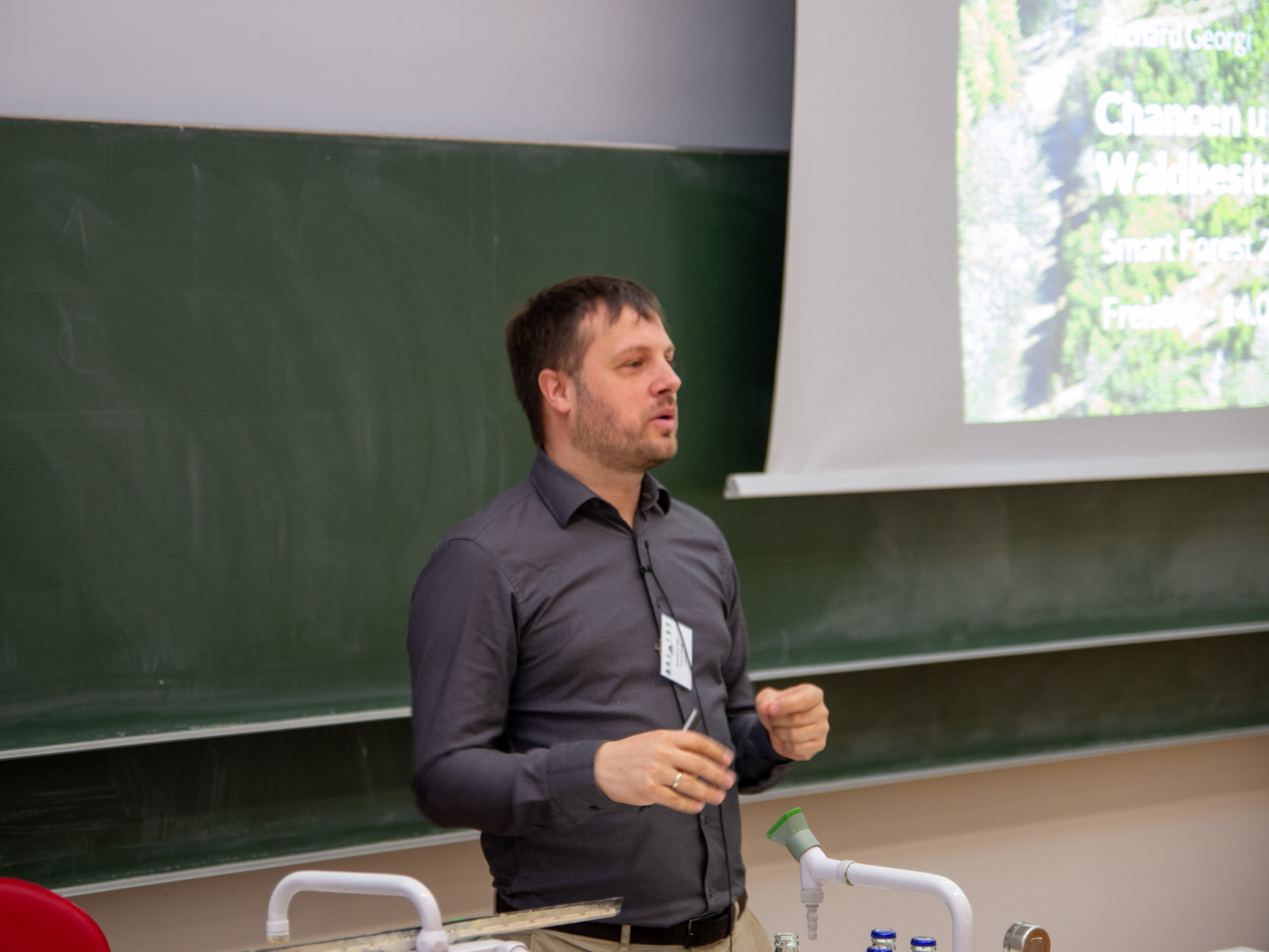 Richard Georgi, a dark-haired man with a stubbly beard, lectures in front of a blackboard and projection screen. On the partially visible screen to the right, we can make out his name, as well as tghe words "Chancen", "Waldbesitz" "Smart Forest" and "Freising".