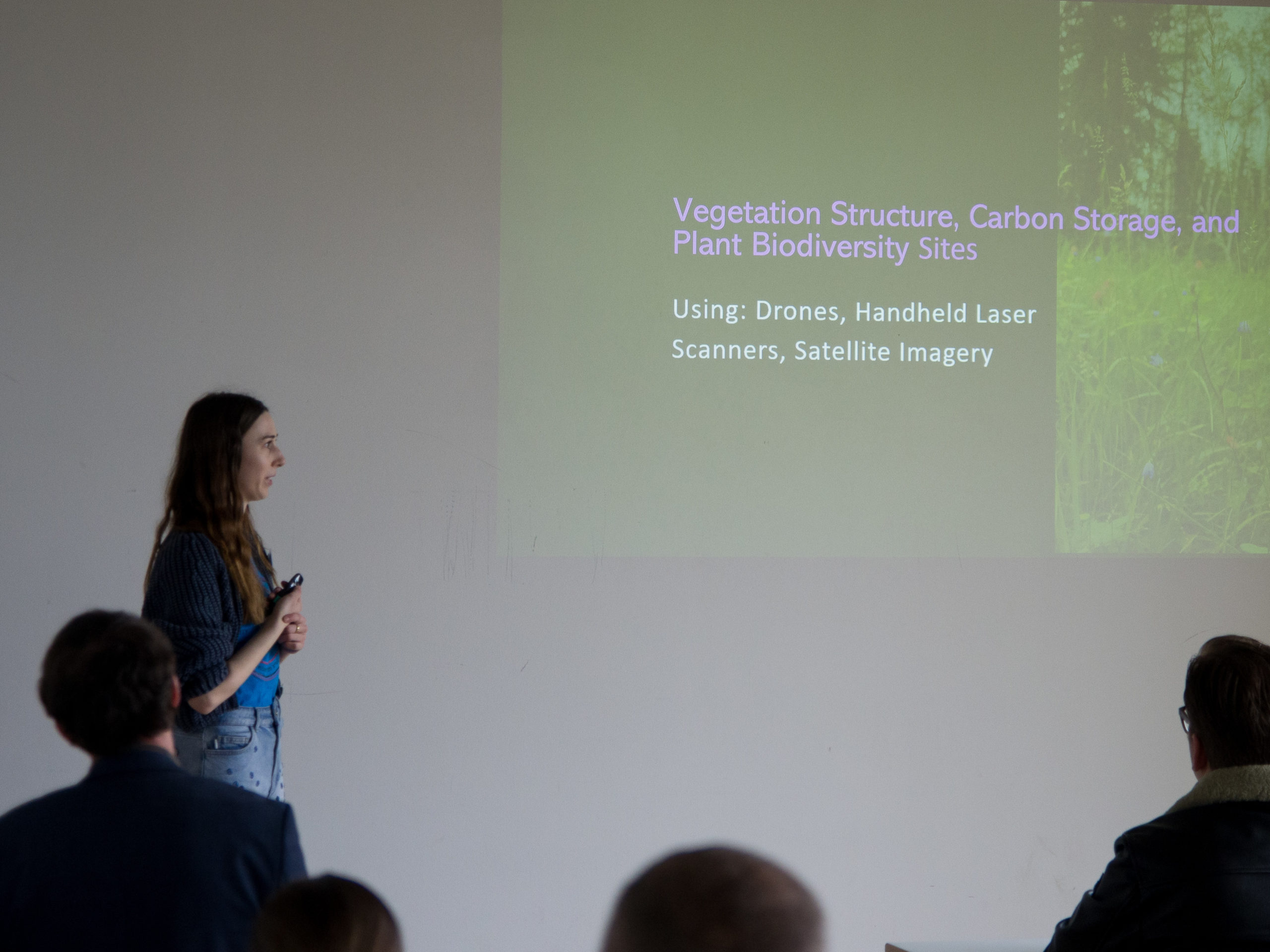 A woman with long brown hair, wearing a blue top and jeans, stands to the left of a projection, explaining something as a crowd seen from the back looks one. The projection reads: "Vegetation Structure, Carbon Storage, and Plant Biodiversity Sites - Using: Drones, Handheld Laser Scanners, Satellite Imagery"