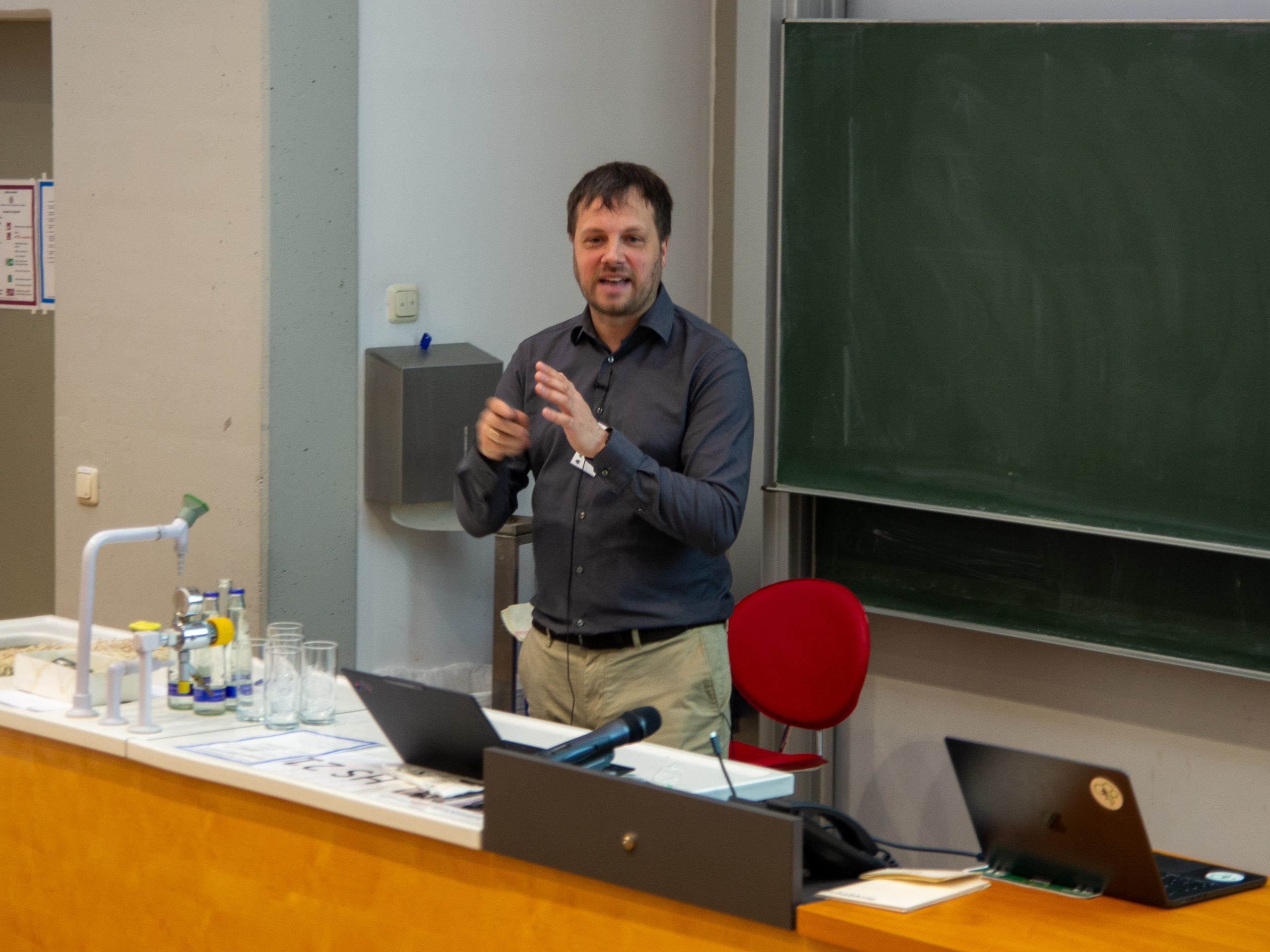 Richard Georgi, a dark-haired man in a charcoal button-down shirt and khaki trousers, lectures from behind a desk. Behind him is a blackboard.