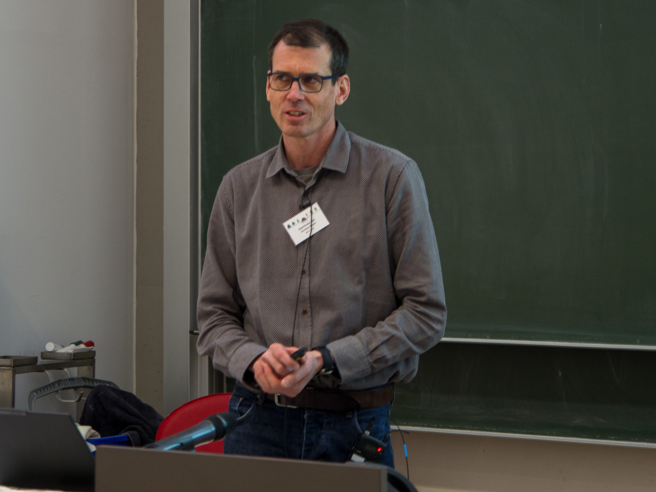 A man in a grey button-up shirt stands in front of a blackboard, his hands clasped in front of him and a microphone visibly hooked up to the front of his shirt. His expression is a thinking one, as if he's in the midst of answering a question.